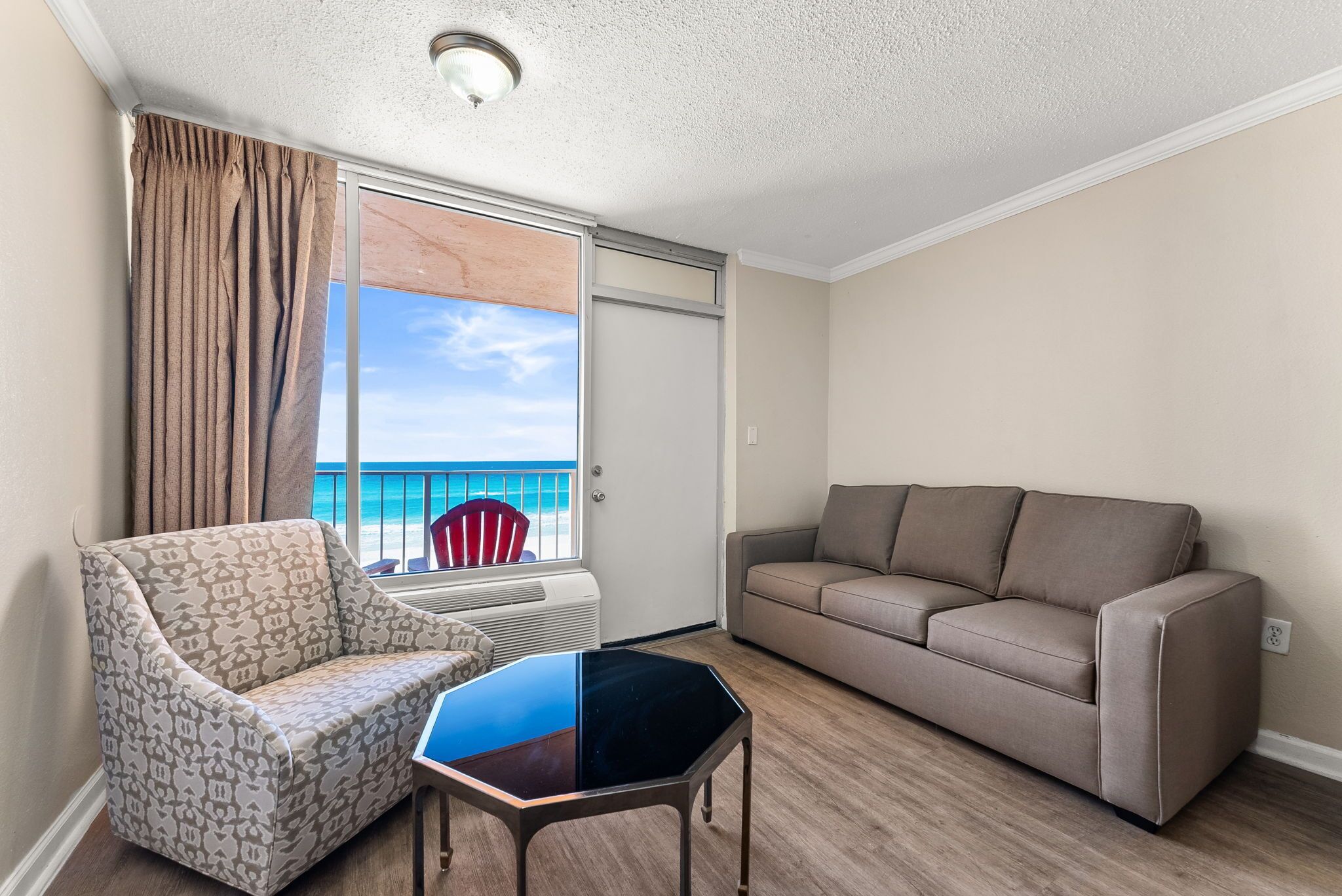 A room with a patterned armchair, a sofa, a small table, and a view of the beach through a sliding door.