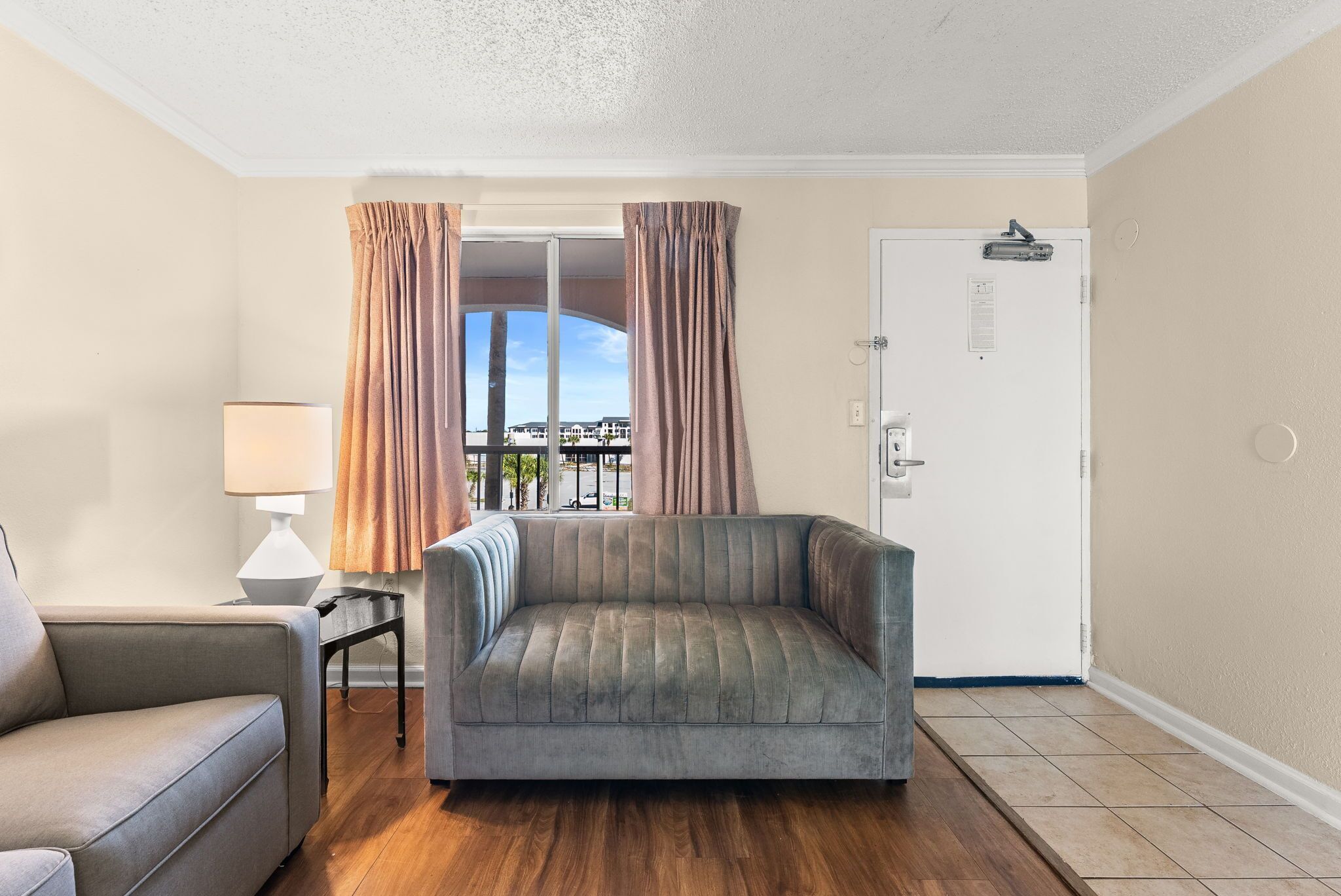 A room with a grey sofa, a smaller armchair, a lamp, and a door with a view of the outside.