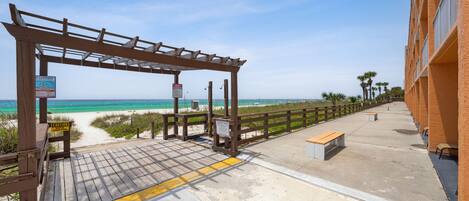 A wooden boardwalk leading to a beach with a wooden pergola, benches, and a building with balconies.