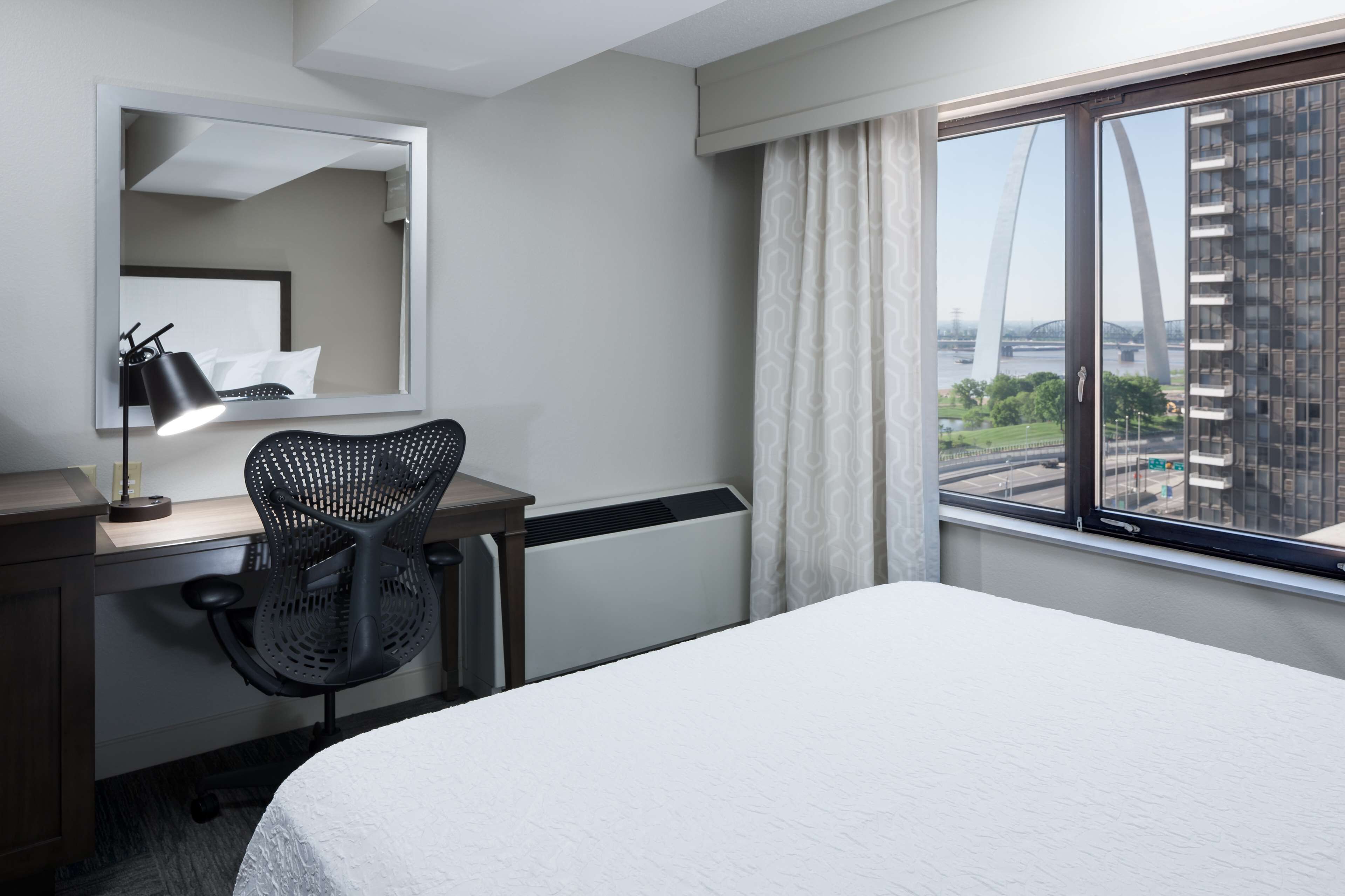 Une chambre d’hôtel comprenant un lit, un bureau avec une chaise, une lampe de bureau, une fenêtre donnant sur un pont et des immeubles, ainsi qu’un grand miroir.