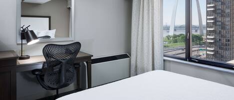 Une chambre d’hôtel comprenant un lit, un bureau avec une chaise, une lampe de bureau, une fenêtre donnant sur un pont et des immeubles, ainsi qu’un grand miroir.