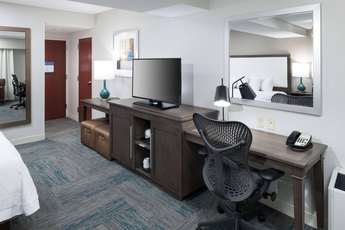 Quarto de hotel com cama, escrivaninha com computador, cadeira, televisão, luminária e telefone.