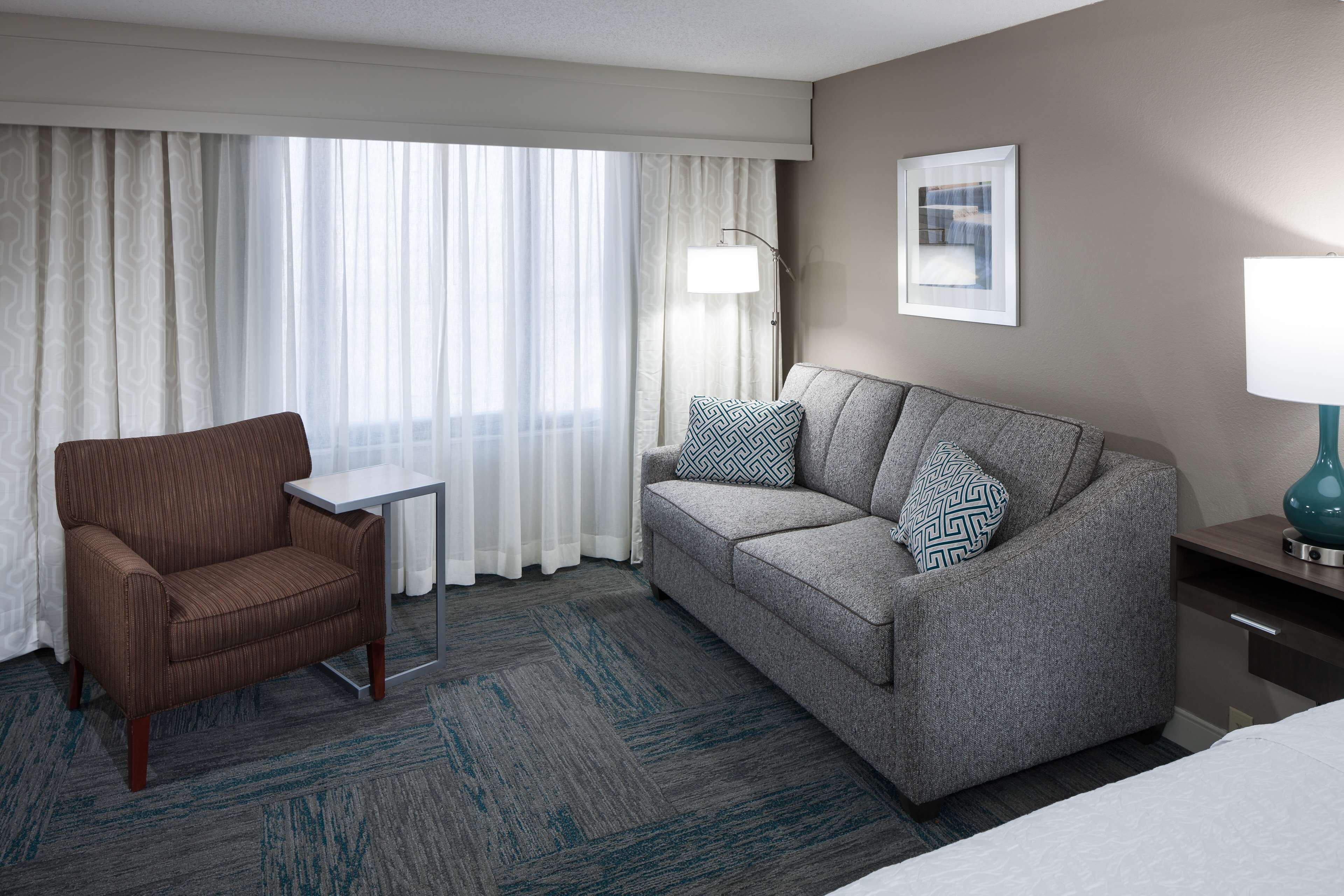 A hotel room with a brown armchair, a grey sofa with patterned cushions, a small table, a lamp, and a framed picture on the wall.