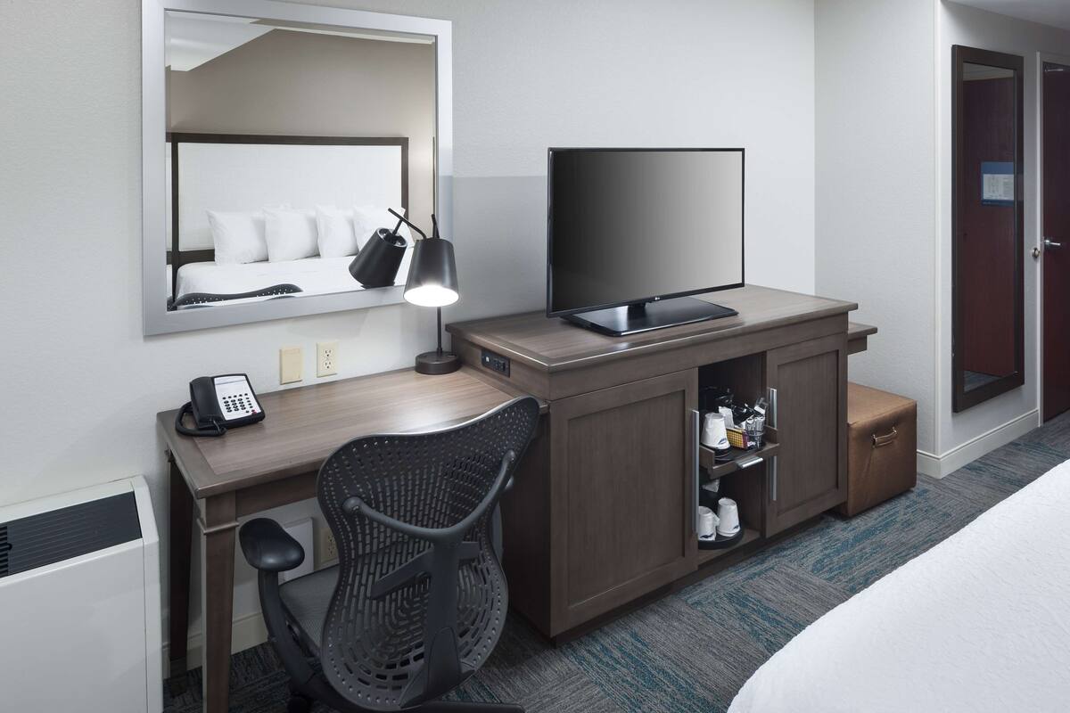Quarto de hotel com cama, escrivaninha com computador, cadeira, televisão, luminária e minibar.