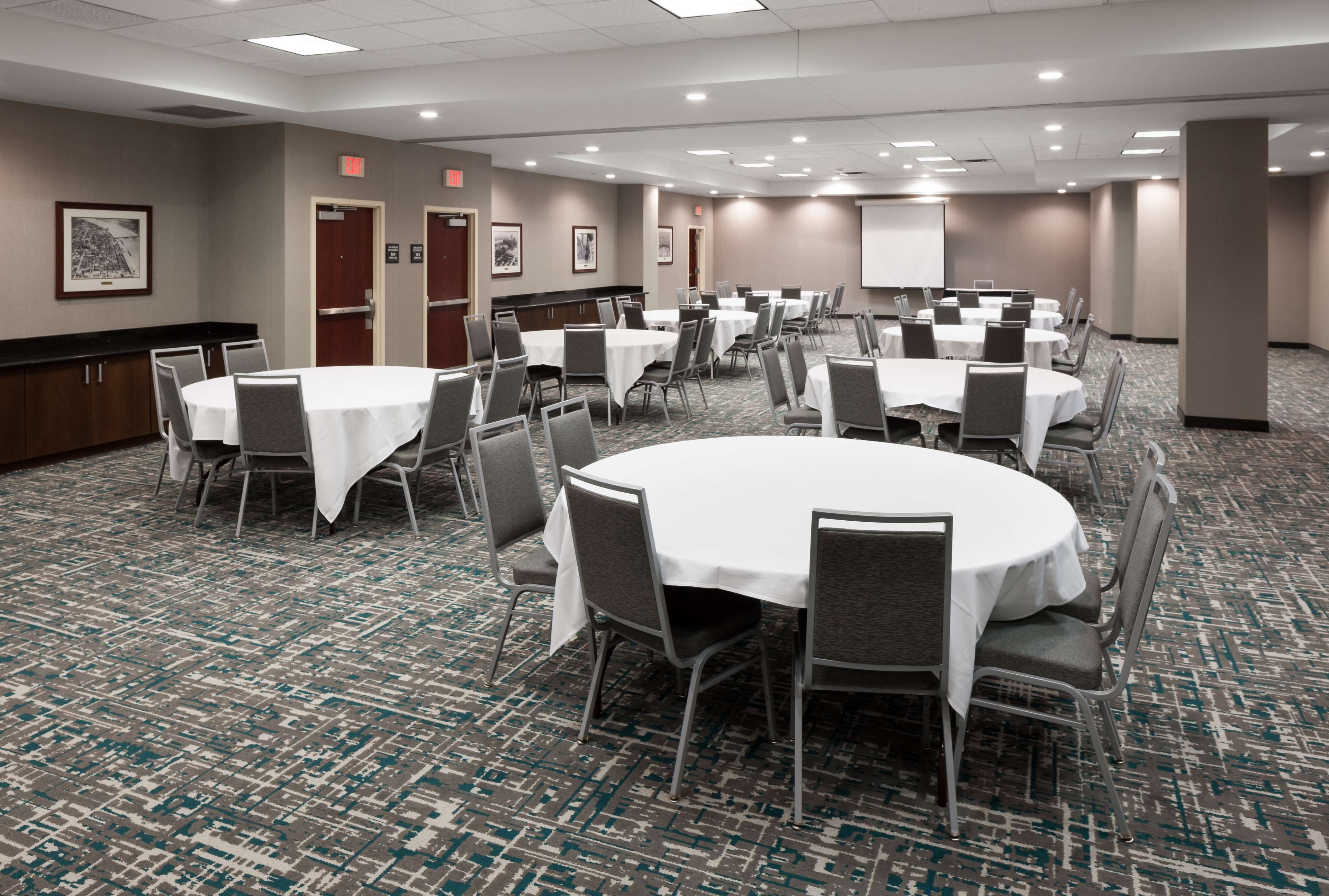 A conference room with multiple round tables, each surrounded by chairs, and a projector screen on the wall.