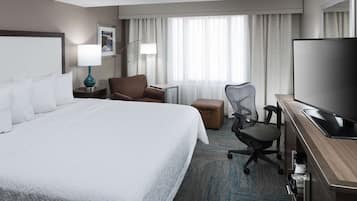 A hotel room with a large bed, a desk with a flat-screen TV, a chair, a lamp, and a window with curtains.