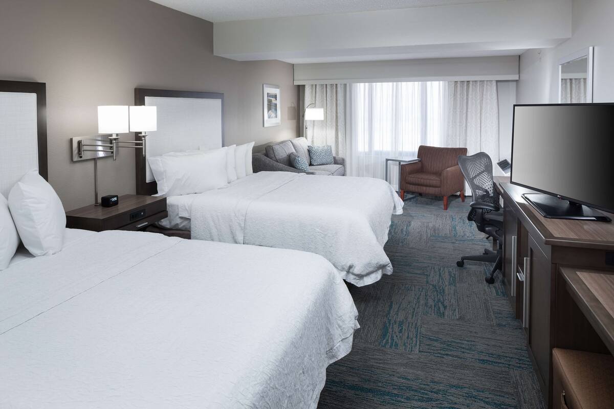 Quarto de hotel com duas camas, uma escrivaninha com televisão, uma cadeira e um abajur.