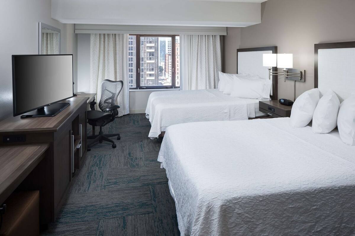Quarto de hotel com duas camas, uma mesa de escritório com televisão, uma cadeira, uma janela com cortinas e uma mesa de cabeceira com abajur.