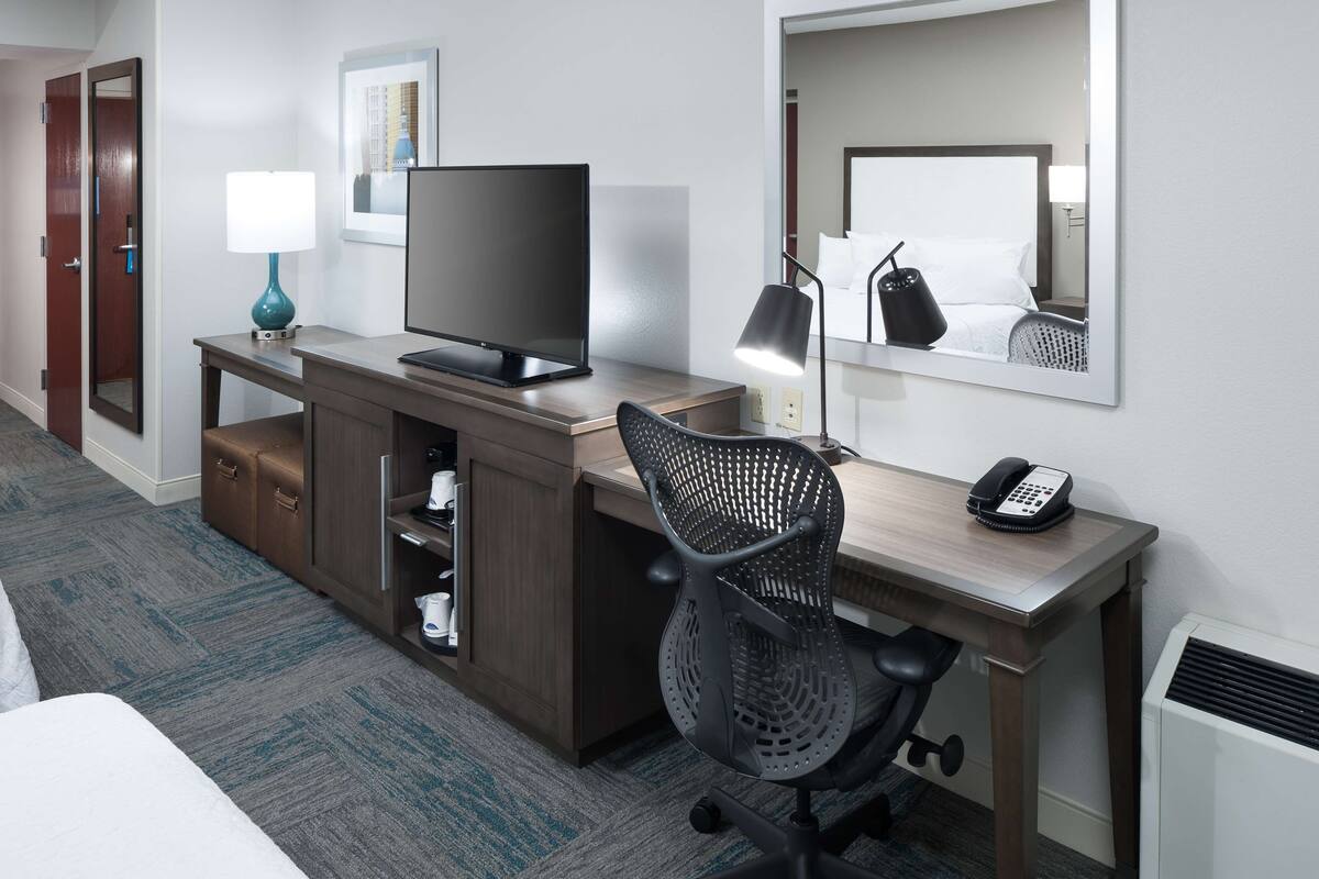 Quarto de hotel com uma escrivaninha de madeira, televisão, cadeira, telefone, abajur e espelho.