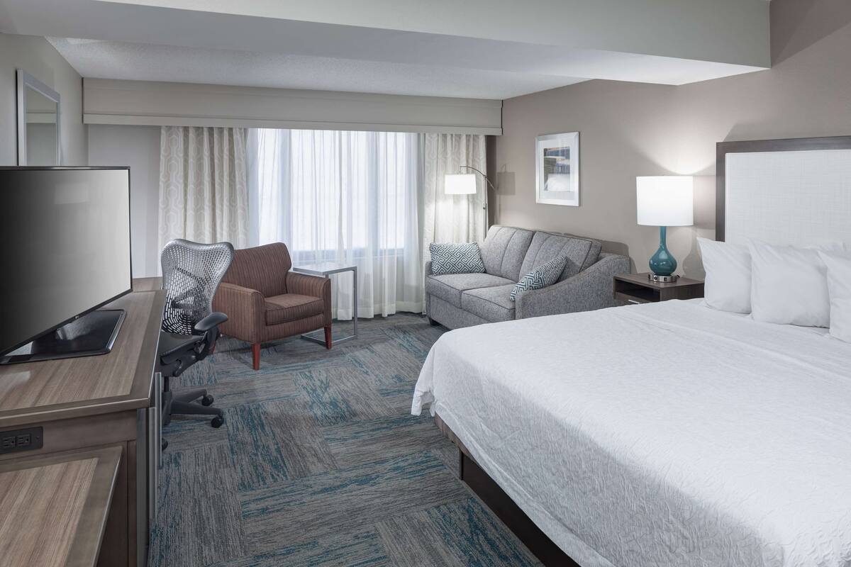 Quarto de hotel com cama, escrivaninha com computador, poltrona, sofá e televisão.