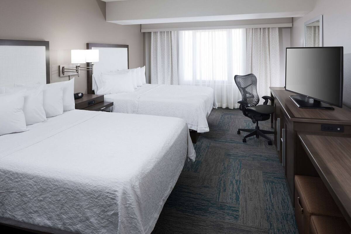 Quarto de hotel com duas camas, uma escrivaninha com televisão, uma cadeira e uma janela com cortinas.