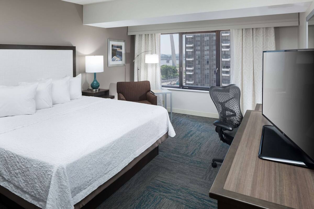 Quarto de hotel com uma cama grande, uma mesa com televisão de tela plana, uma cadeira e vista para prédios pela janela.