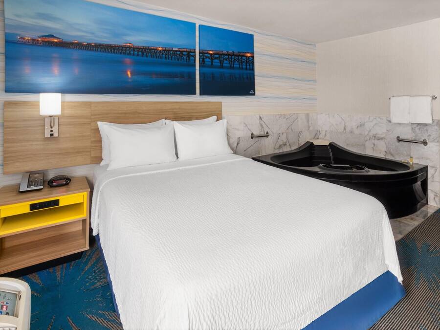 Una habitación de hotel con una cama, una mesita de noche, un escritorio, una lámpara de escritorio, un teléfono, una bañera y una obra de arte colgada en la pared.