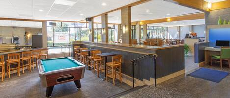 A pool table in a spacious indoor area with wooden chairs and tables, a bar area, and large windows allowing natural light.