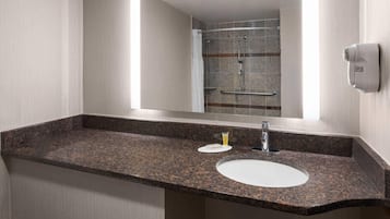 A bathroom with a granite countertop, a white sink, a handheld showerhead, and a towel dispenser.