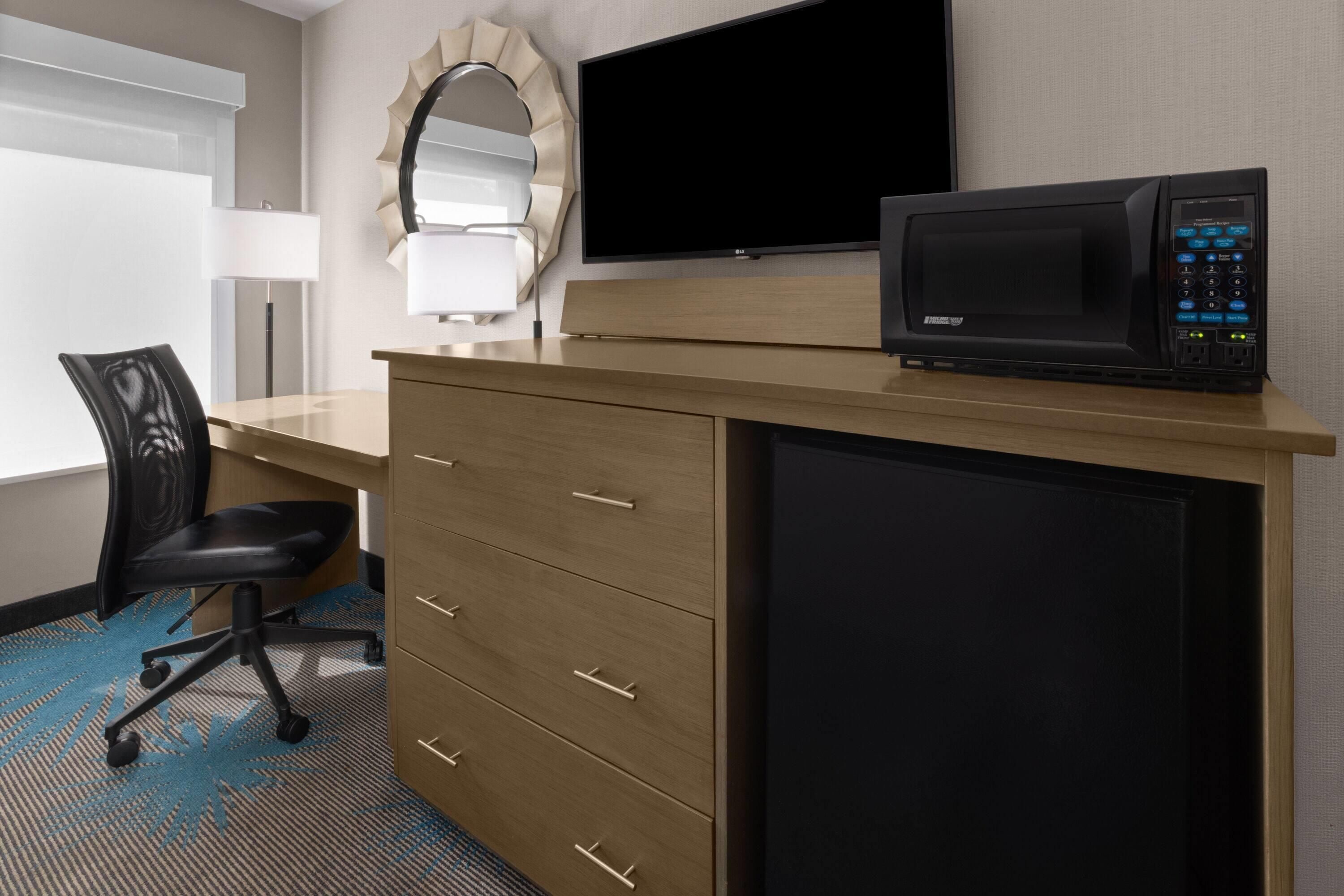 A hotel room with a desk, a black office chair, a flat-screen TV, a microwave, and a large mirror.