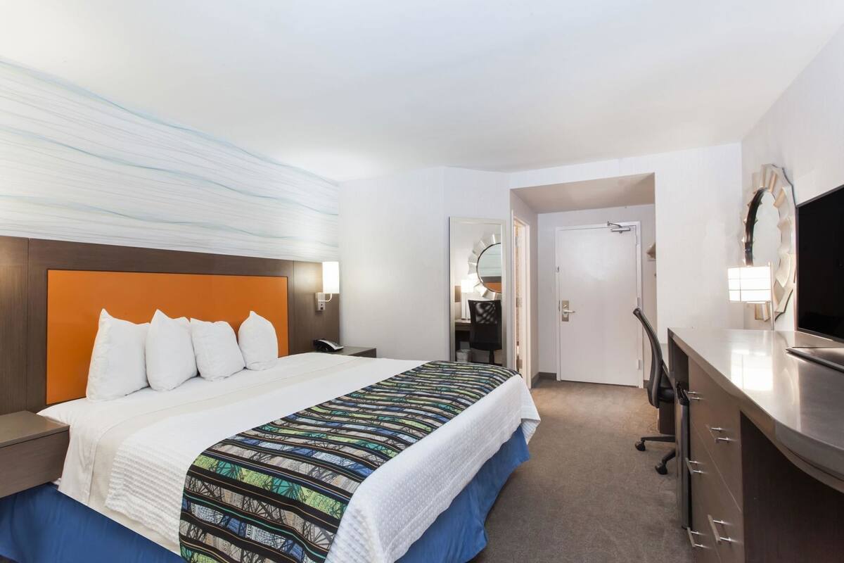 Habitación de hotel con cama, mesita de noche, escritorio, silla, televisor y puerta.