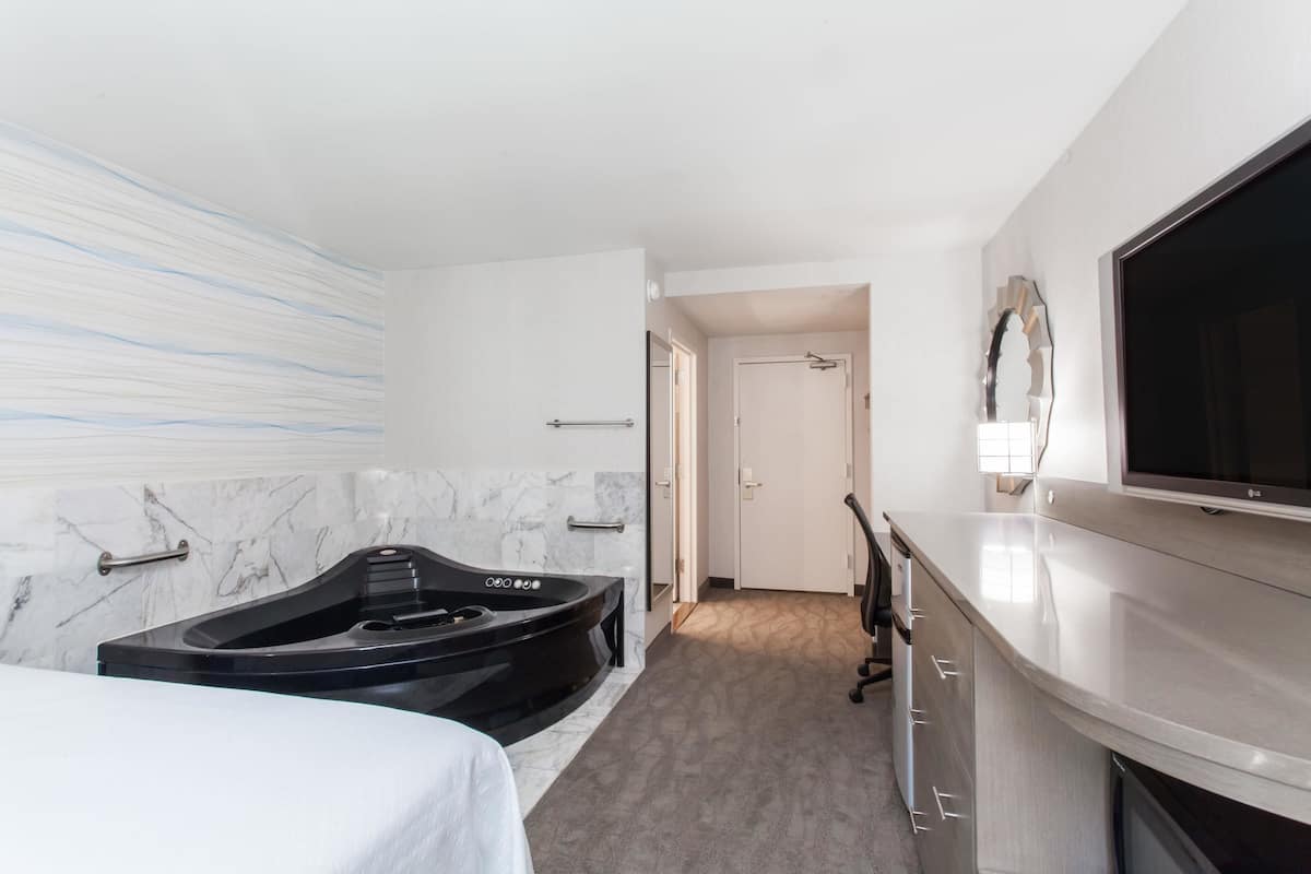 Una habitación de hotel moderna con un jacuzzi negro grande, un televisor de pantalla plana montado en la pared y una pared con diseño de mármol.