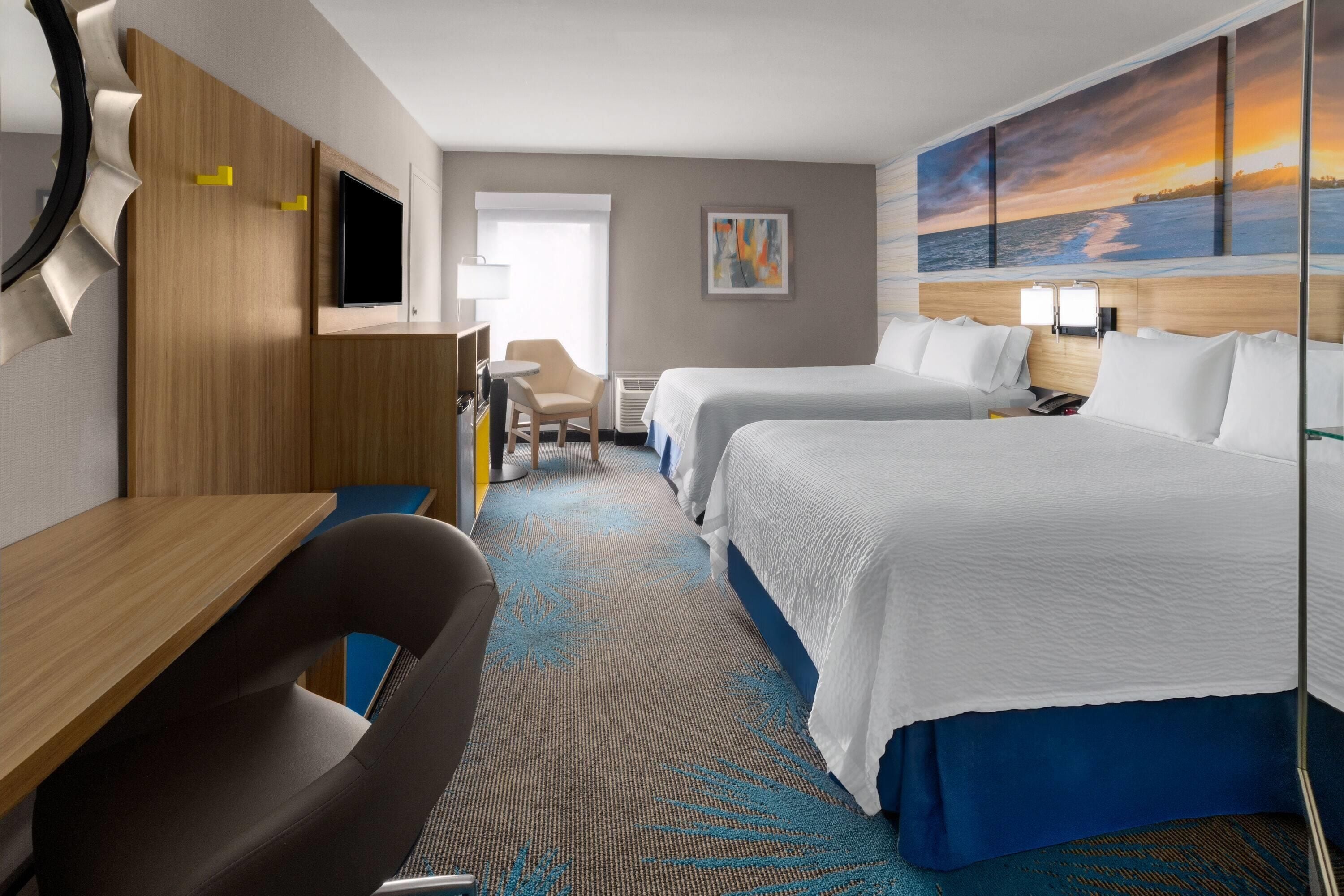 A hotel room with two beds, a desk, a chair, a TV, and a painting of a sunset.