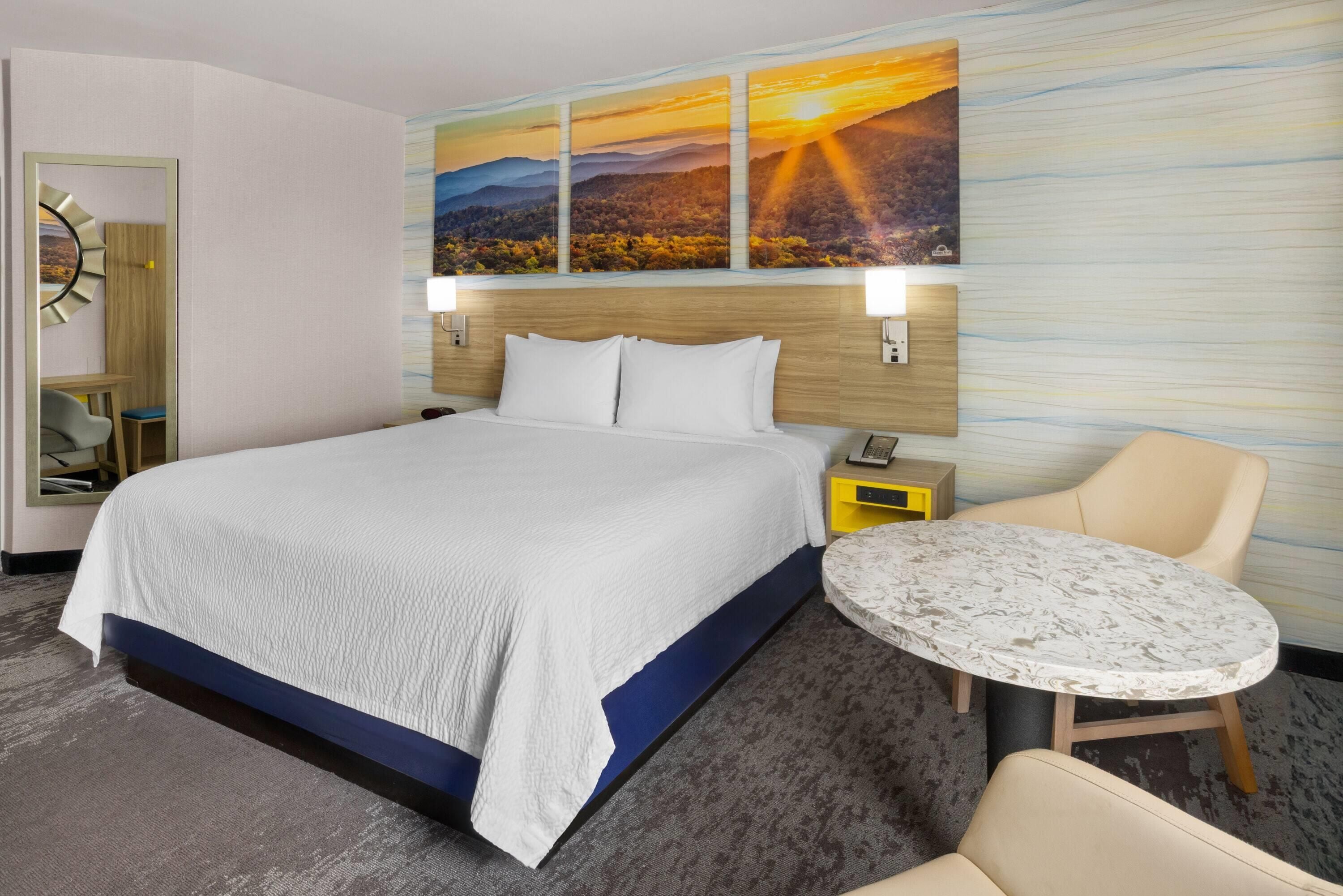 A hotel room with a large bed, a bedside table with a phone, a chair, a small round table, and a wall with a landscape painting.