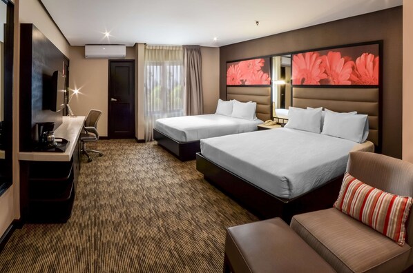 Habitación de hotel con dos camas, un escritorio con una silla, un sofá y un cuadro grande con motivos florales en la pared.