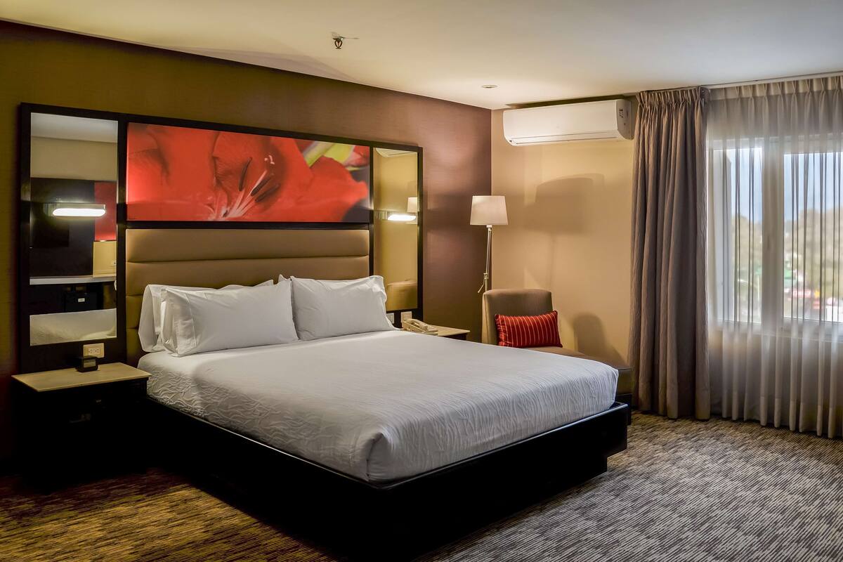 Hotelkamer met een groot bed, nachtkastje, lamp, stoel en een raam met gordijnen.