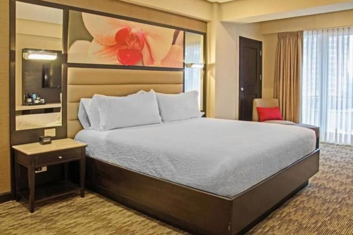 Una habitación de hotel con una cama grande, una mesita de noche, un espejo y una ventana con cortinas.