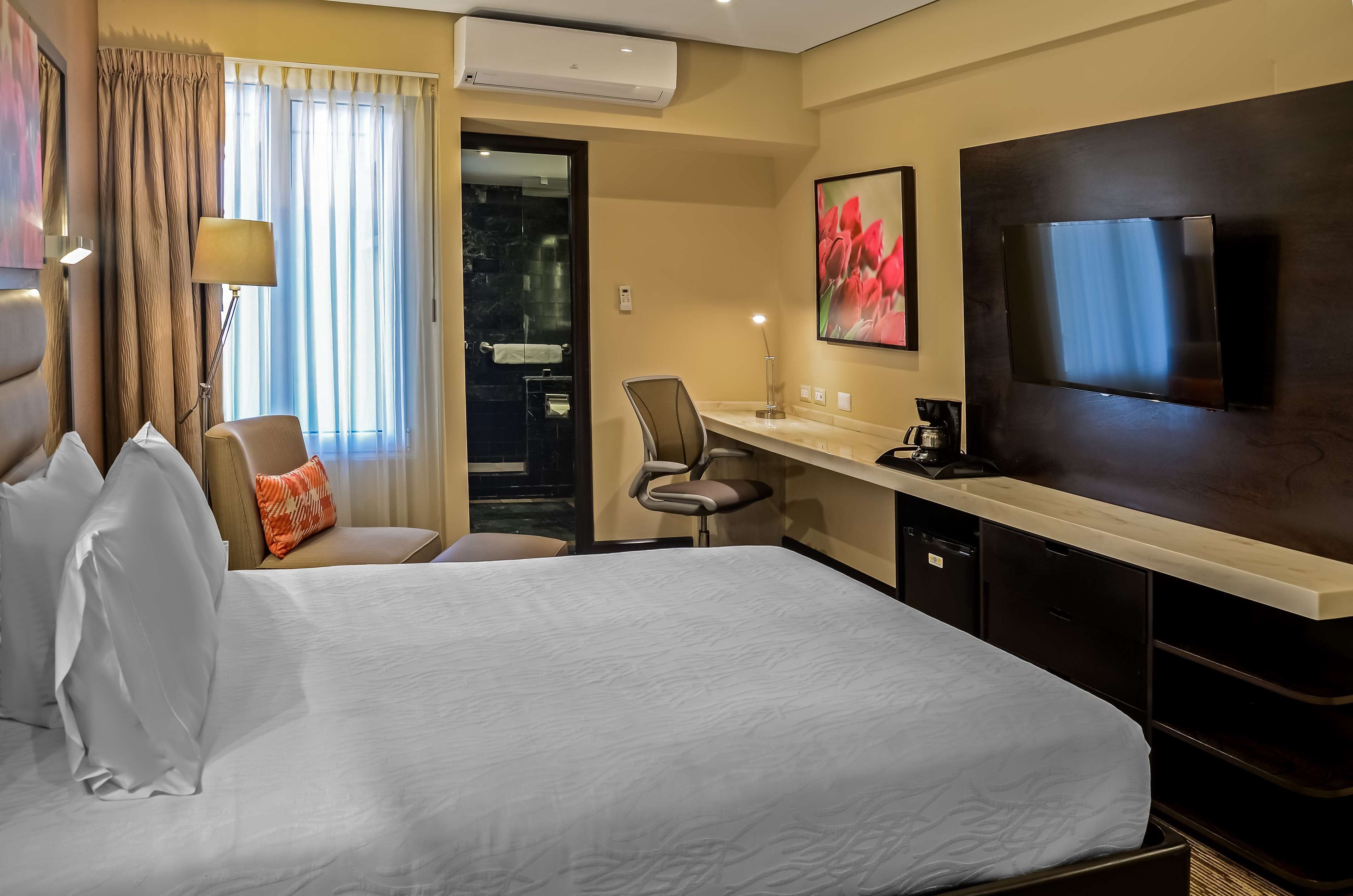 Habitación de hotel con cama, escritorio, silla, televisor y un cuadro.