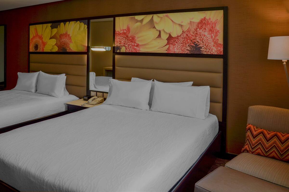 Een hotelkamer met twee bedden, een nachtkastje met telefoon, een stoel en een groot bloemig kunstwerk aan de muur.