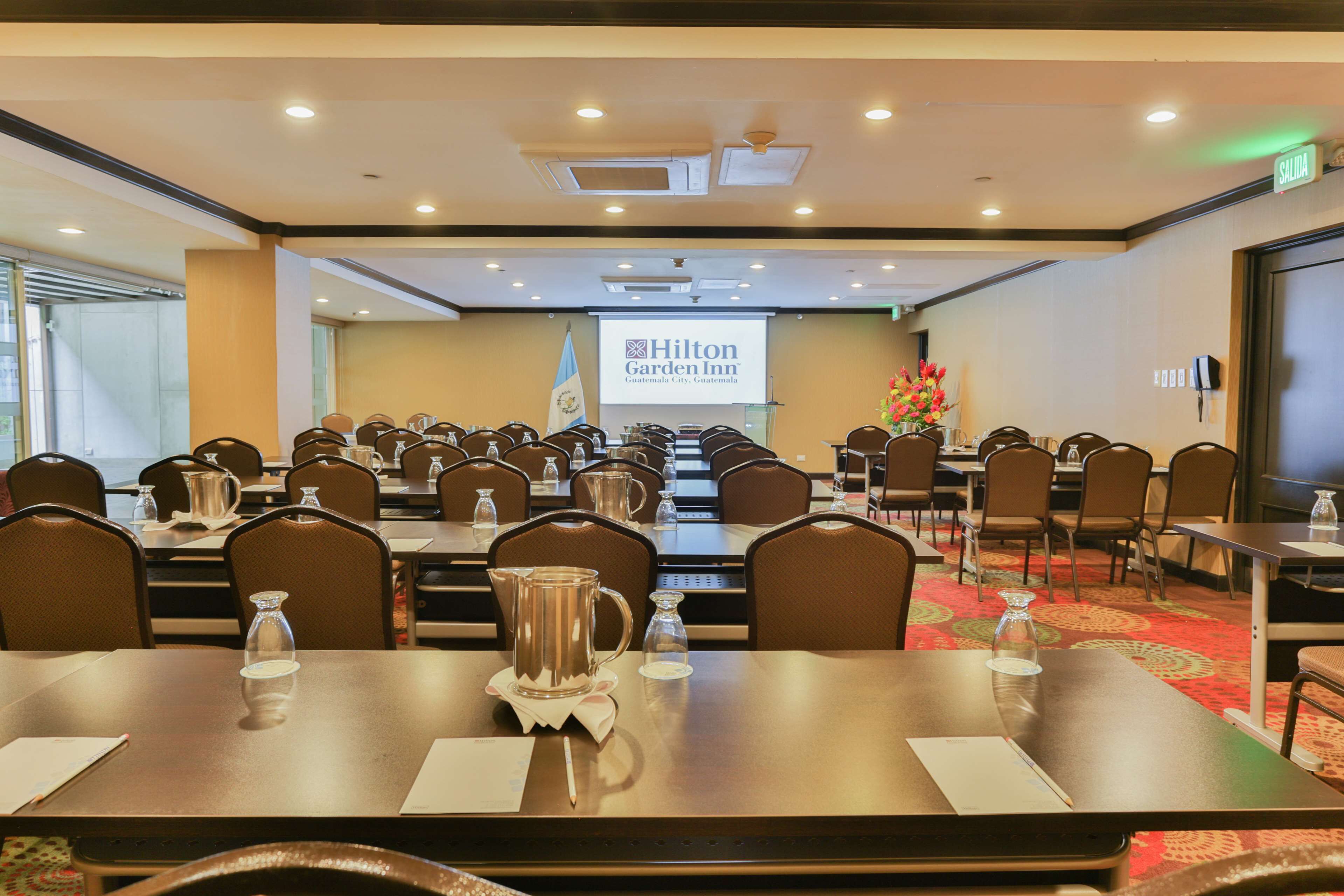 Uma sala de conferências com várias mesas e cadeiras, uma tela de projetor exibindo o logotipo do Hilton Garden Inn e a bandeira de Guatemala.