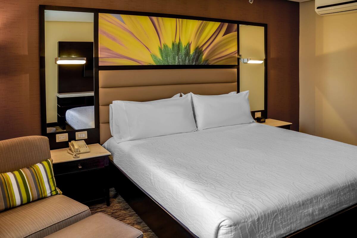 Hotelkamer met een groot bed, een nachtkastje met een telefoon, een stoel met een gestreept kussen, en een kunstwerk aan de muur.