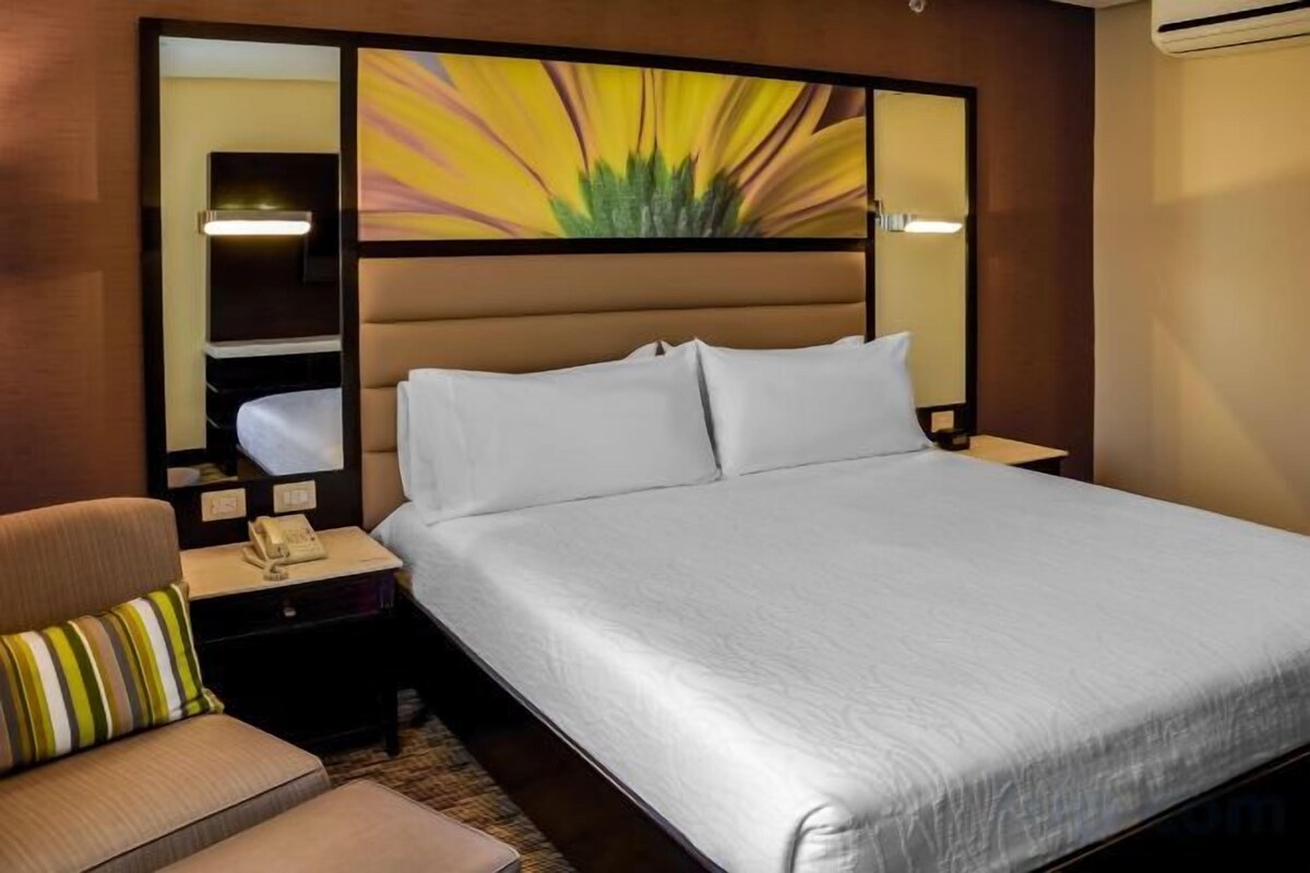 A hotel room with a large bed, a bedside table with a lamp, a chair, and a decorative wall panel.