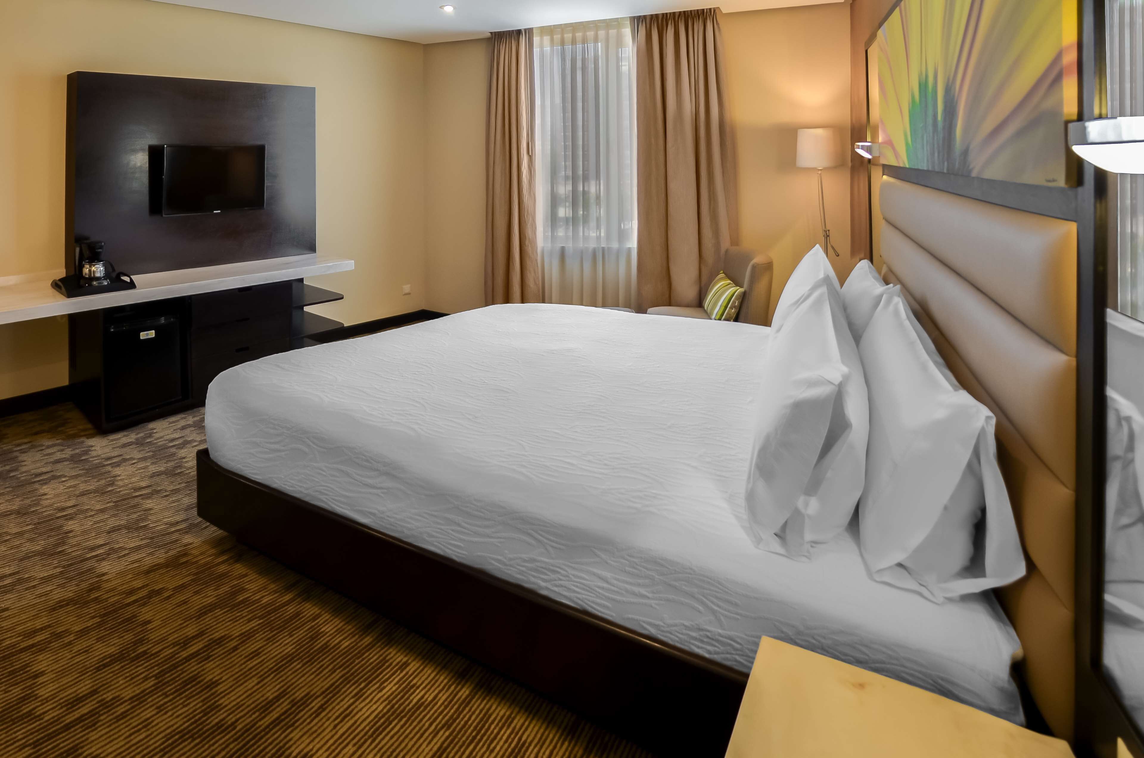 Camera d'albergo con un letto grande, una TV a schermo piatto montata a parete, una scrivania con una macchina per caffè e una finestra con tende leggere.