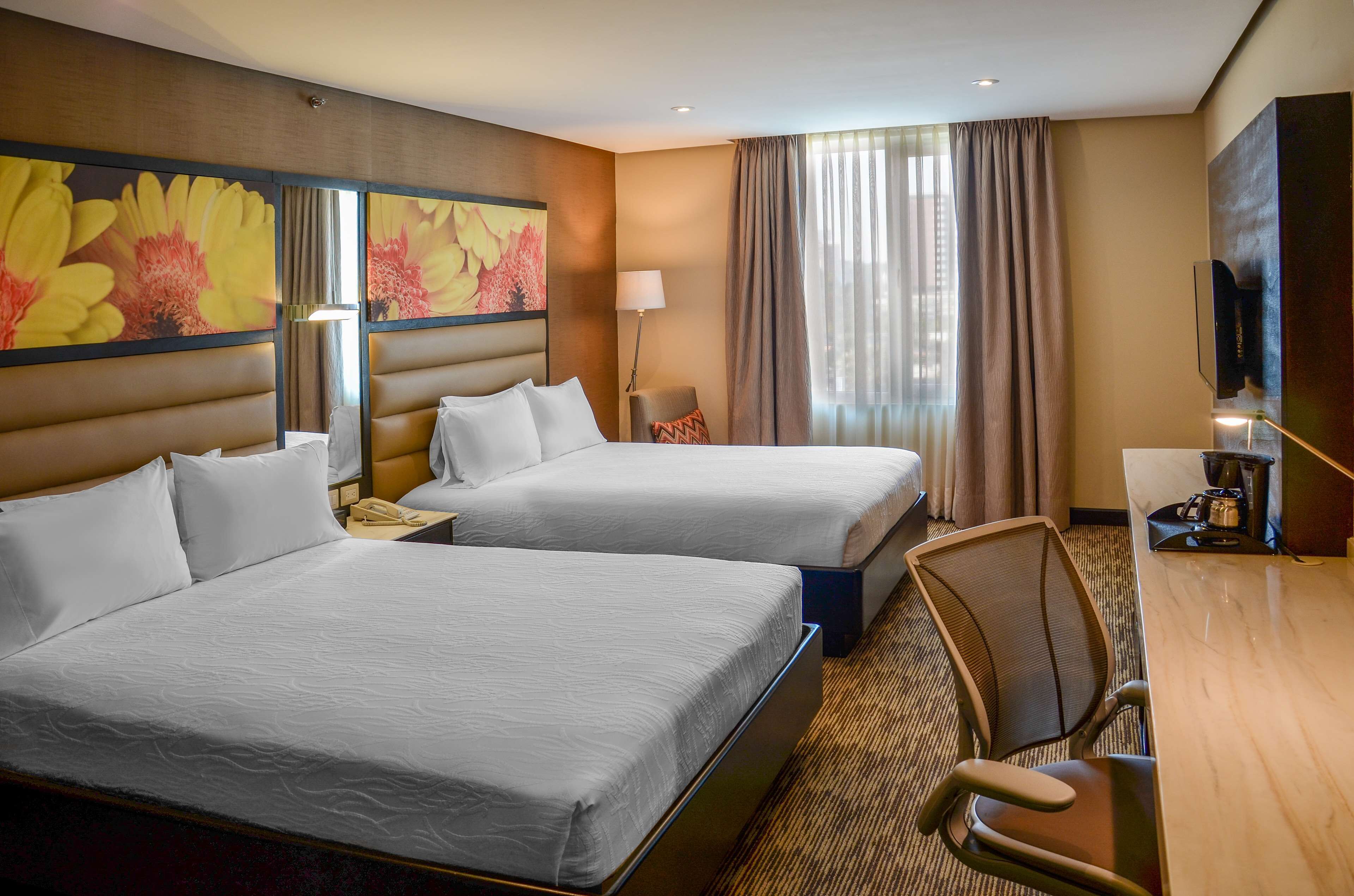 A hotel room with two beds, a desk, a chair, a TV, and a large floral artwork on the wall.
