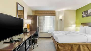 Habitación de hotel con cama, escritorio con televisor, silla, espejo, ventana con cortinas e imagen en la pared.