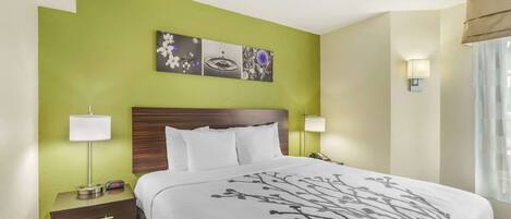 Une chambre d’hôtel comprenant un lit, deux tables de chevet avec des lampes, une table d’appoint, une œuvre d’art accrochée au mur et une fenêtre avec des rideaux.