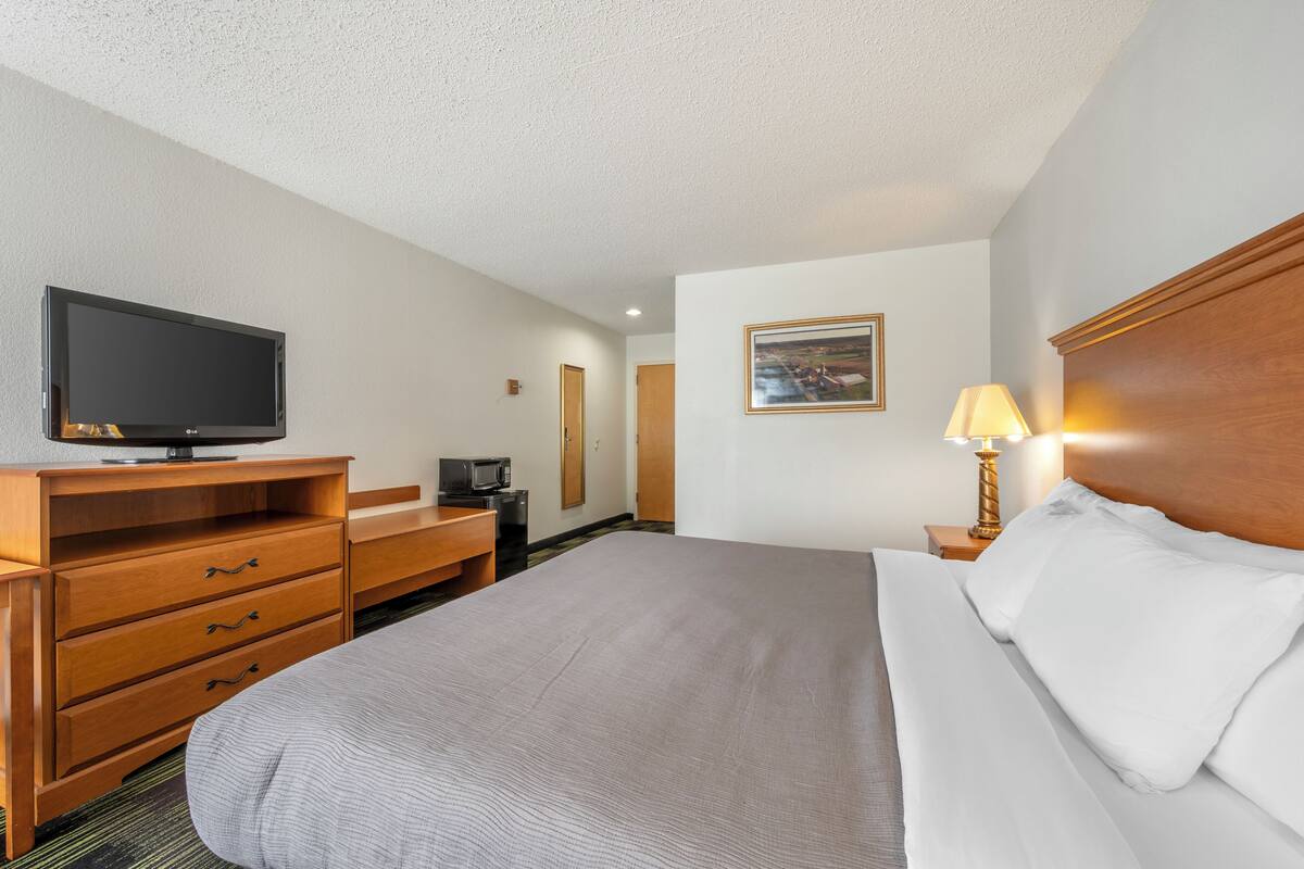 A hotel room with a large bed, a television on a wooden dresser, a desk with a computer, and a lamp.