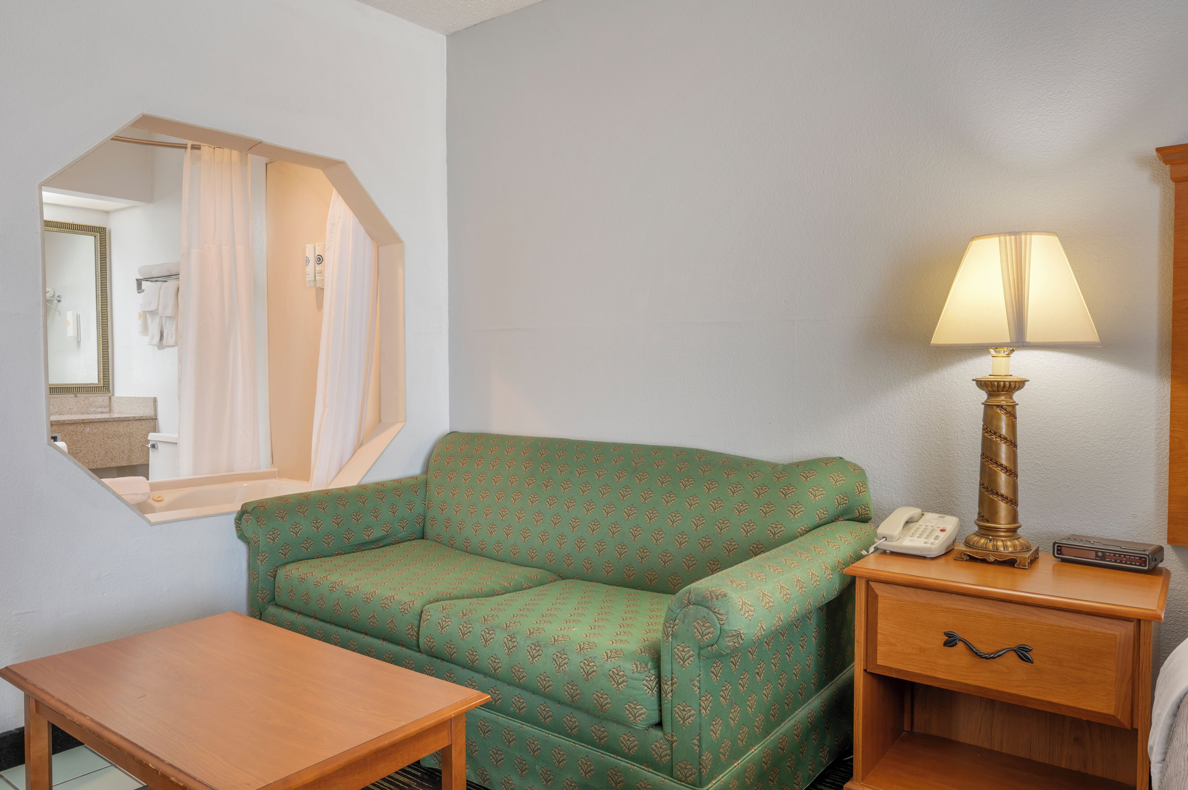 A room with a green sofa, a wooden nightstand with a lamp, a telephone, and a mirror.