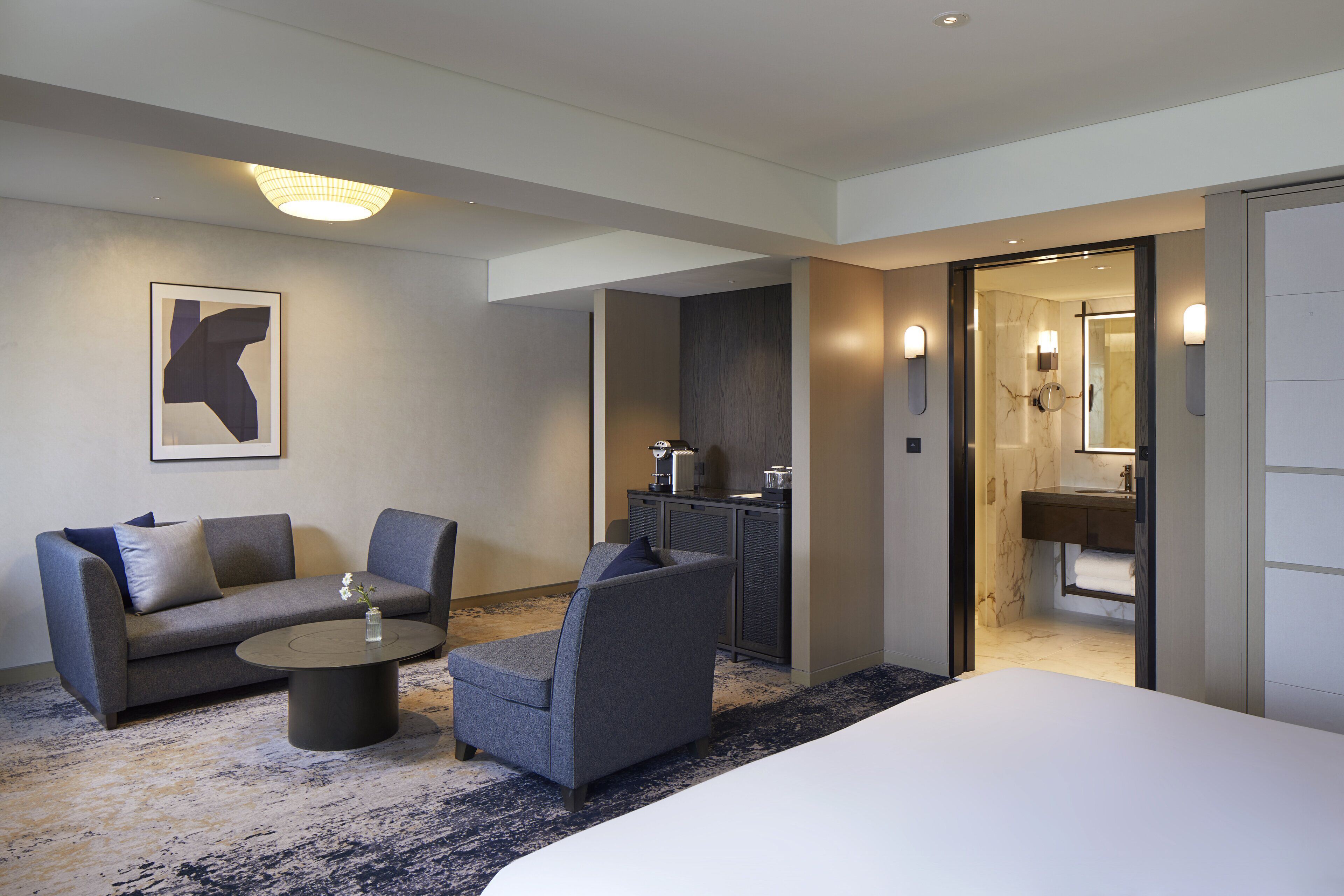Una habitación de hotel moderna con una cama grande, un sofá, dos sillones, una mesa de centro y un pequeño escritorio con lámpara.