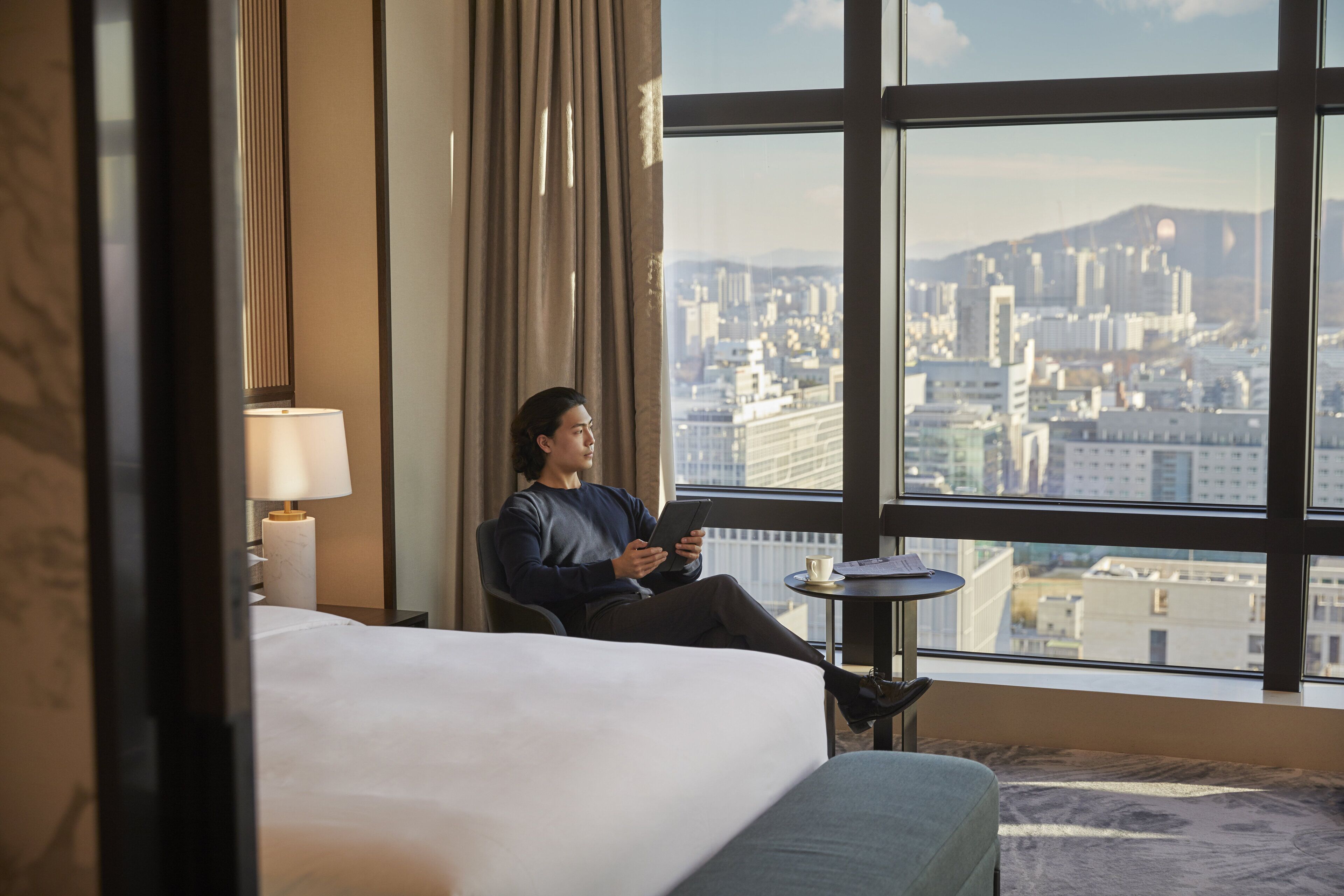 A hotel room with a bed, a bedside table with a lamp, a small round table, and a view of a cityscape through large windows.