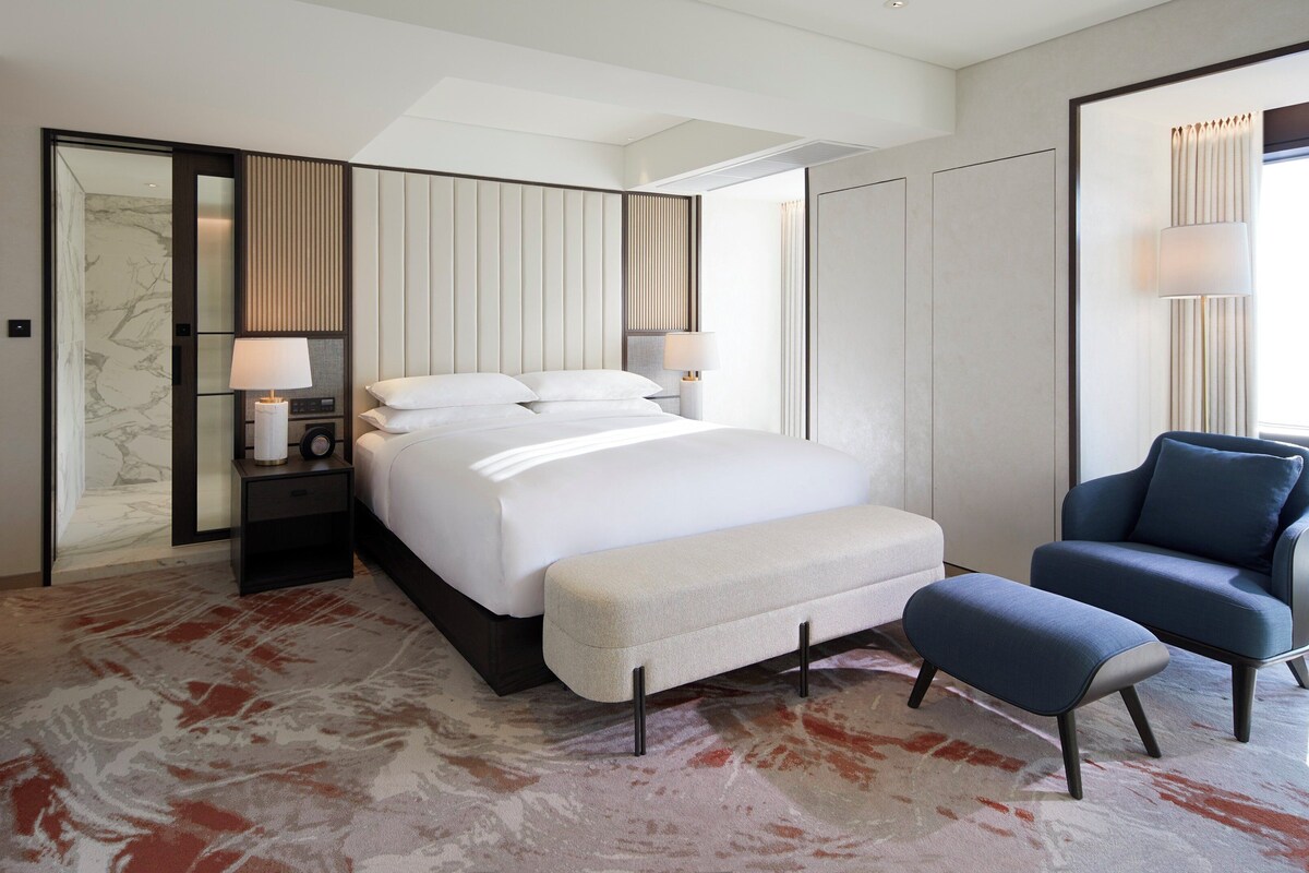 A modern hotel room with a large bed, bedside lamps, a chair, and a view of the city.