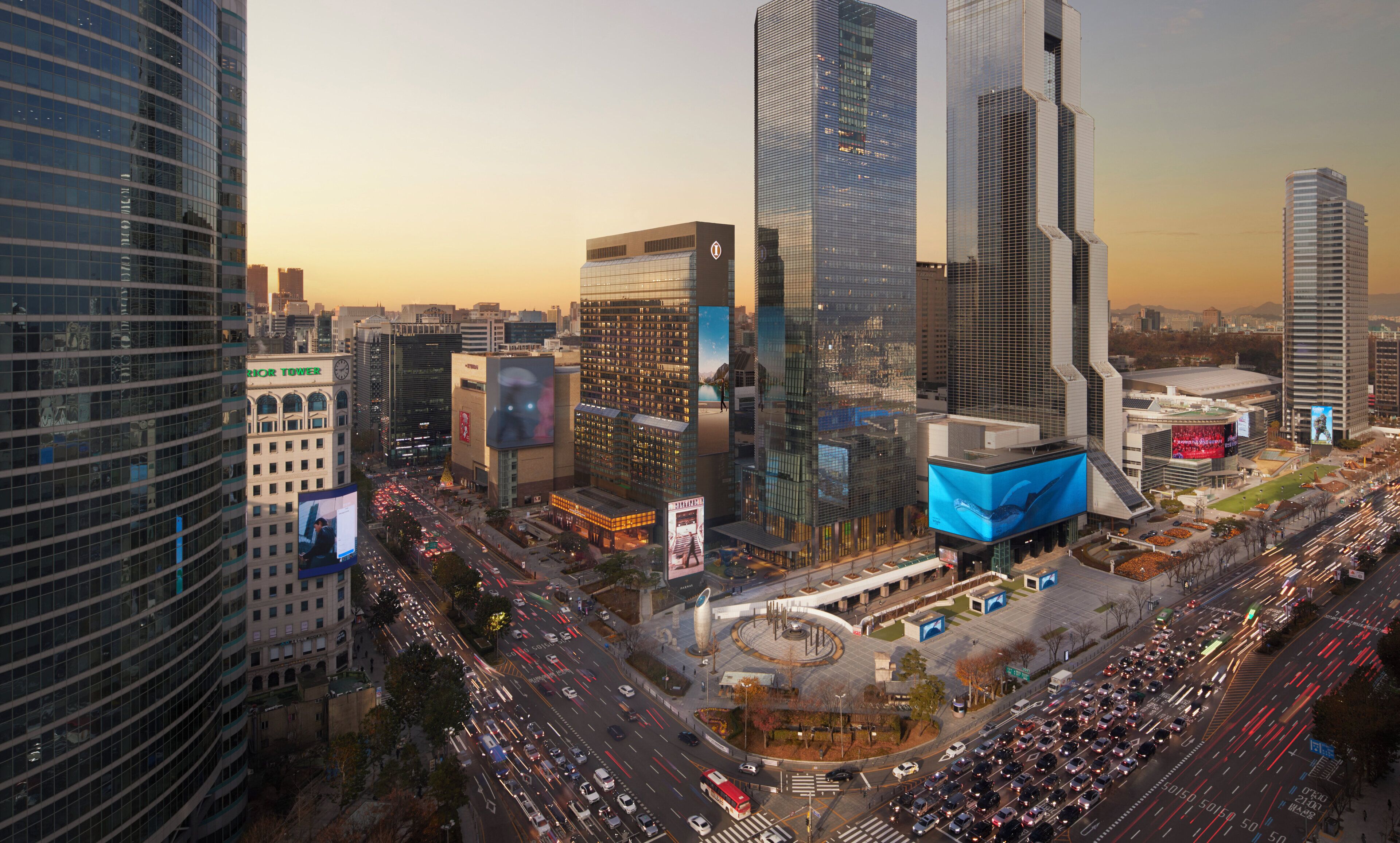 Panorama urbano con moderni grattacieli, un incrocio trafficato e un grande cartellone pubblicitario.