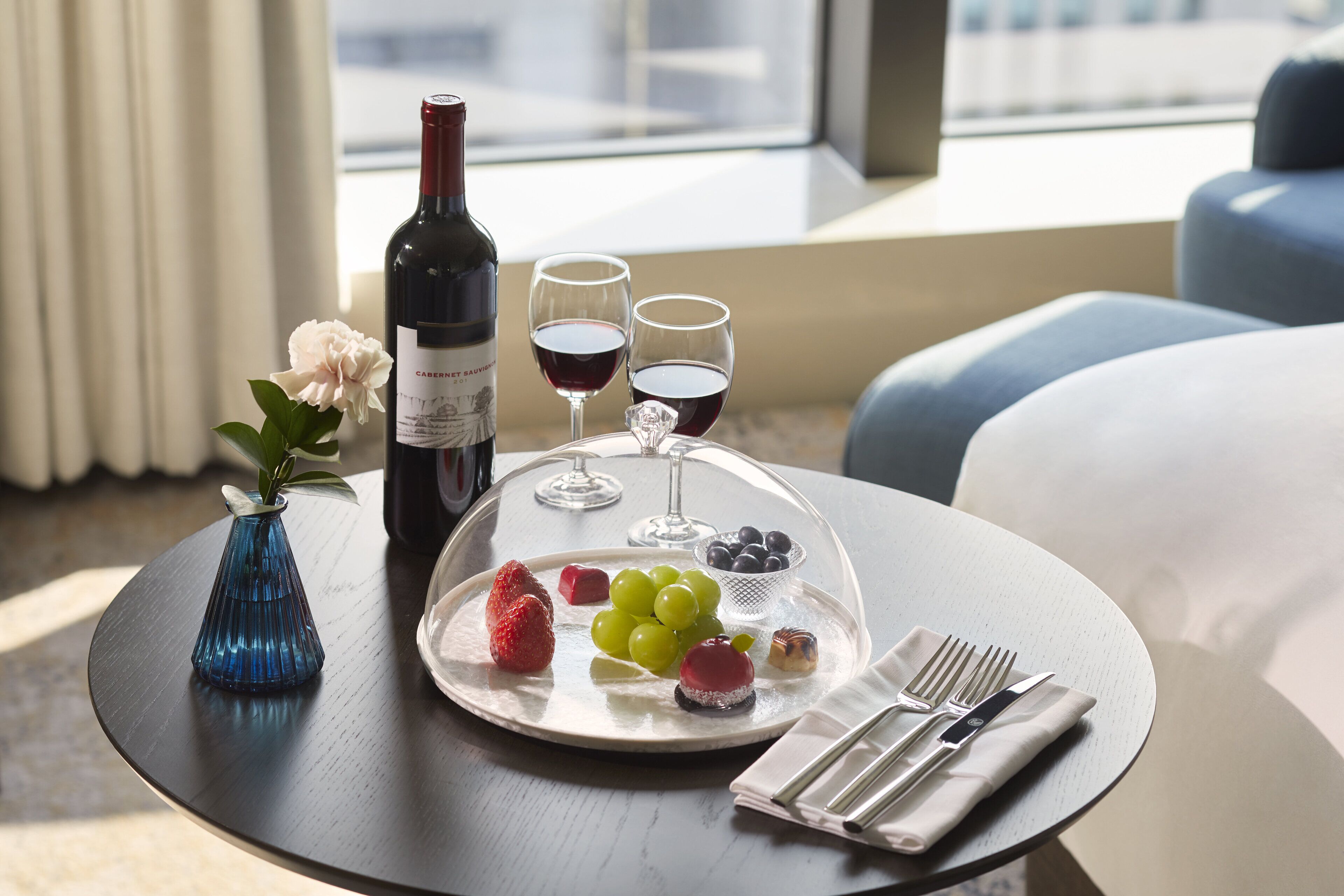 A table with a wine bottle, two glasses of red wine, a plate with grapes, strawberries, and a small dessert, a vase with a flower, and a napkin with cutlery.