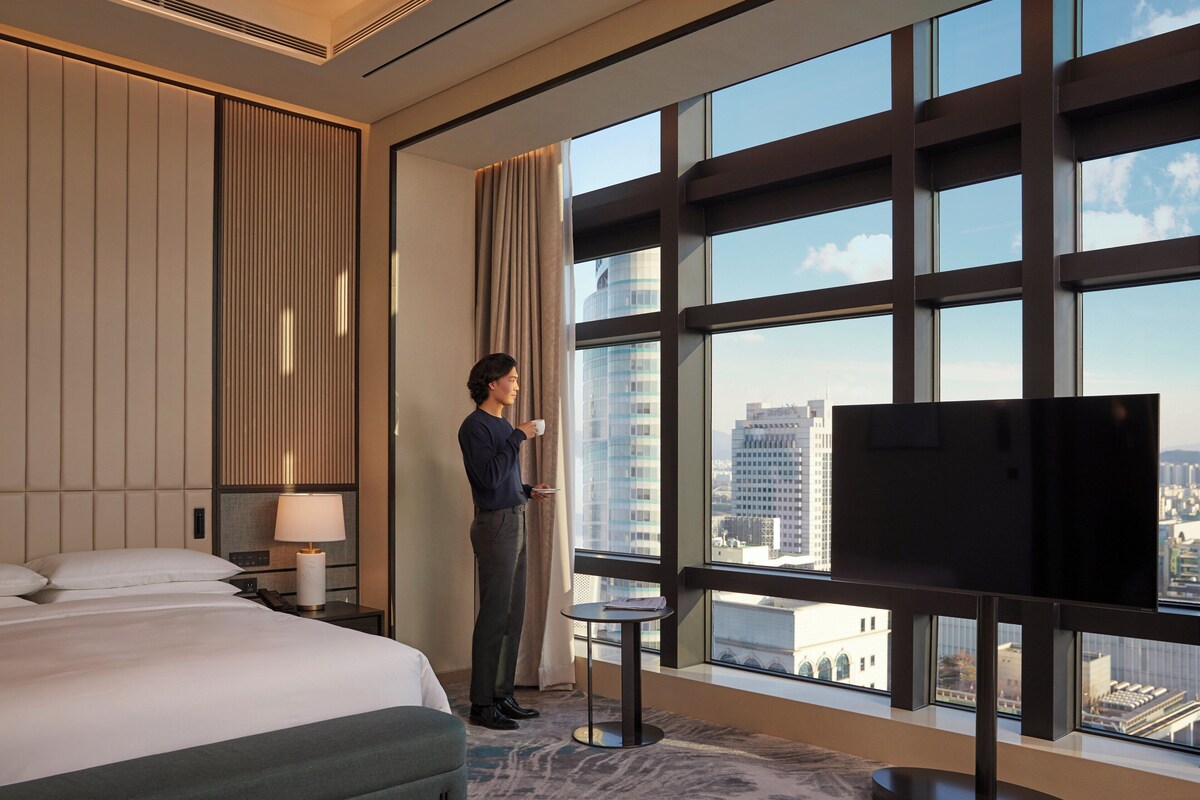 A hotel room with a large bed, a small table, a TV, and a view of the city through large windows.