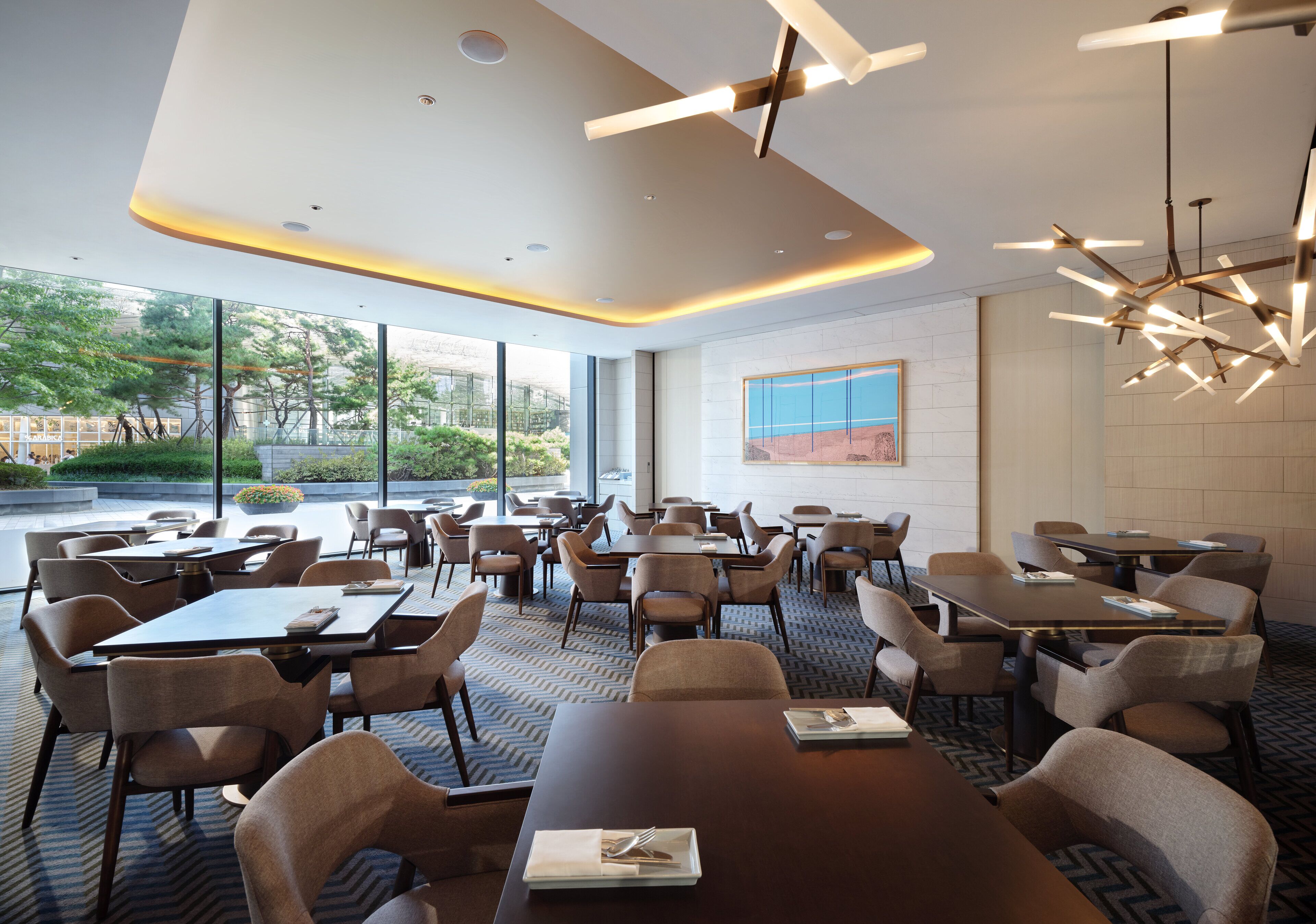 A modern hotel dining area with tables and chairs, large windows, and a view of greenery.