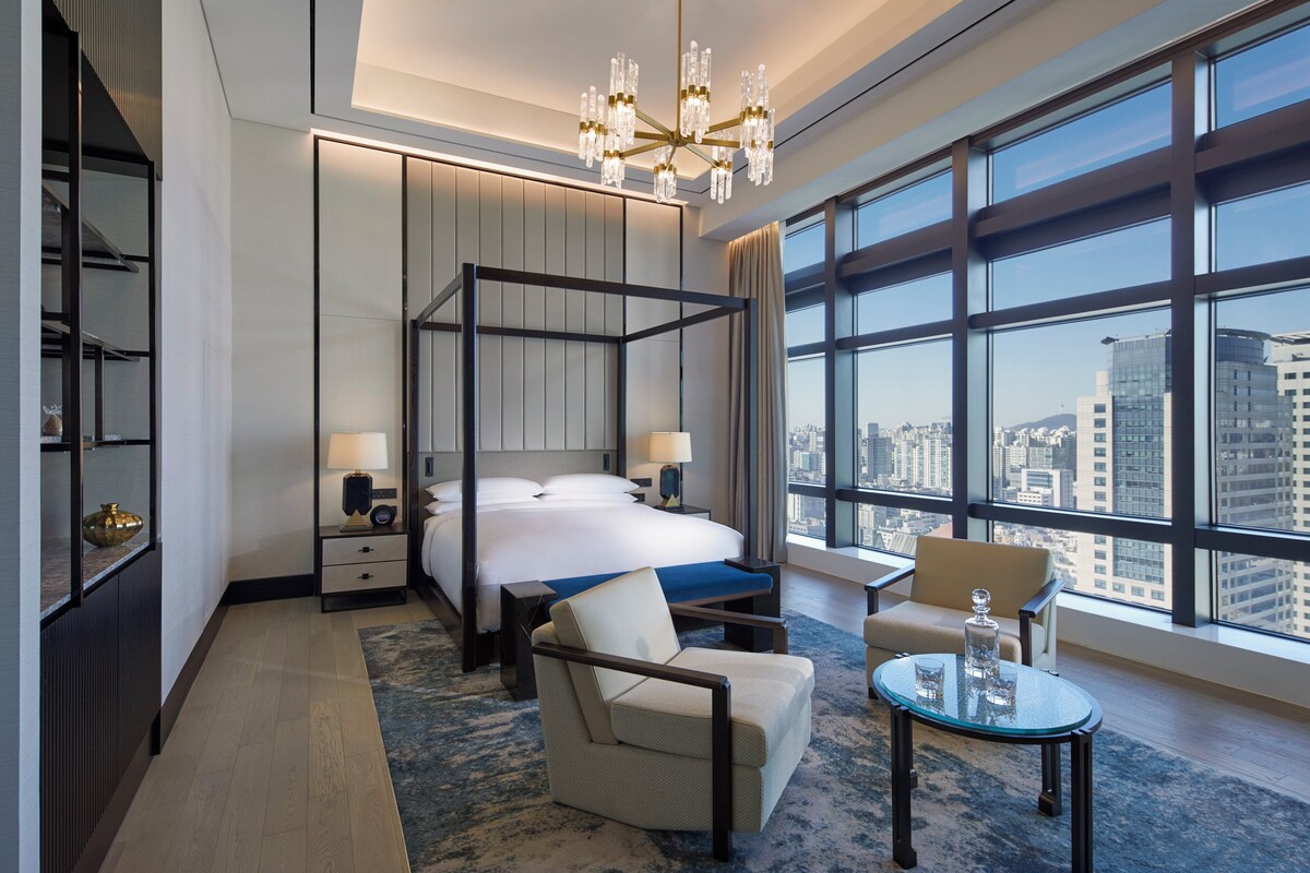 A modern hotel room with a four-poster bed, a sitting area with a chair and table, and large windows offering a city view.