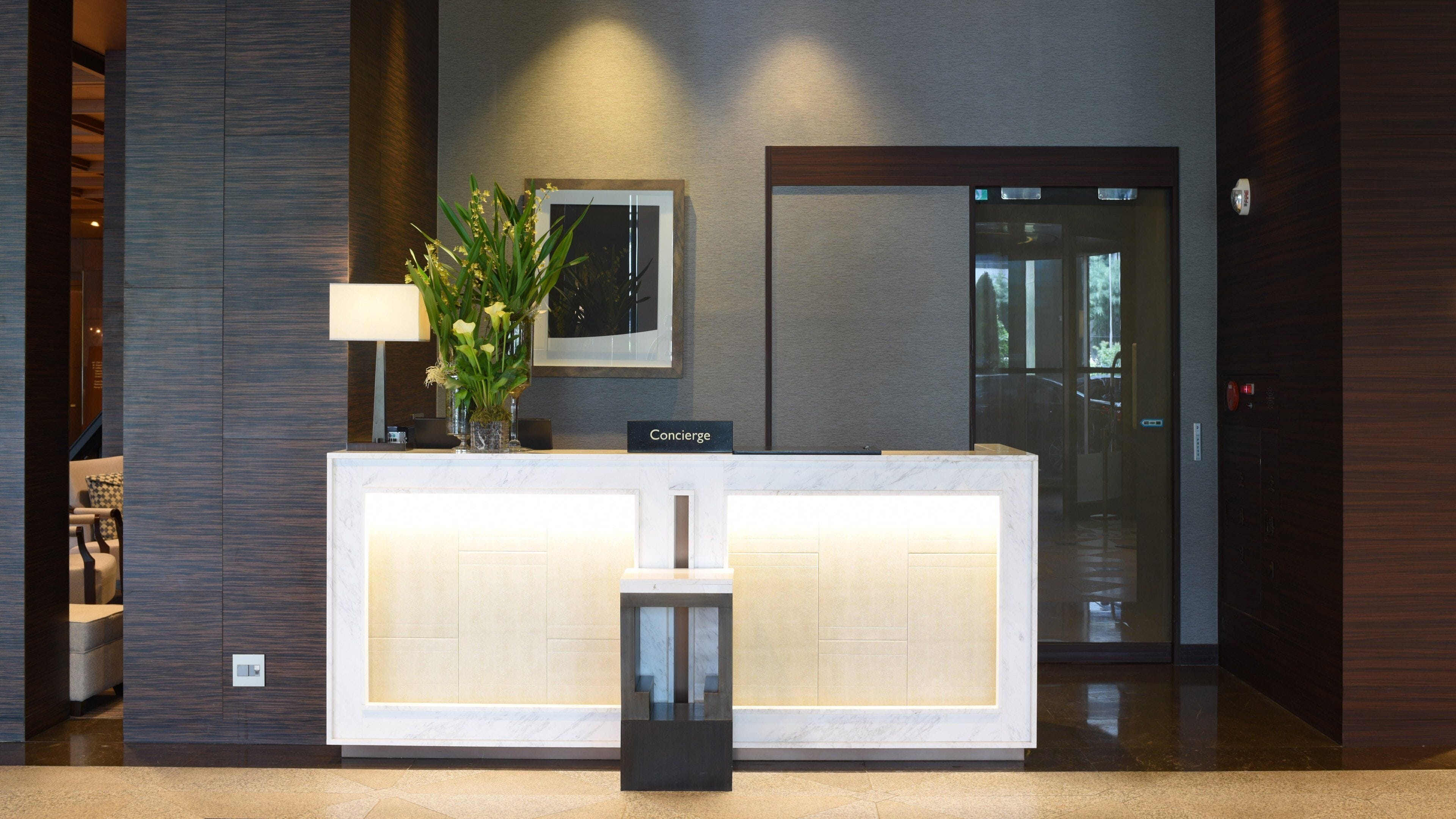 A reception desk with a sign reading "Concierge," a vase with flowers, a lamp, and a framed picture on the wall.