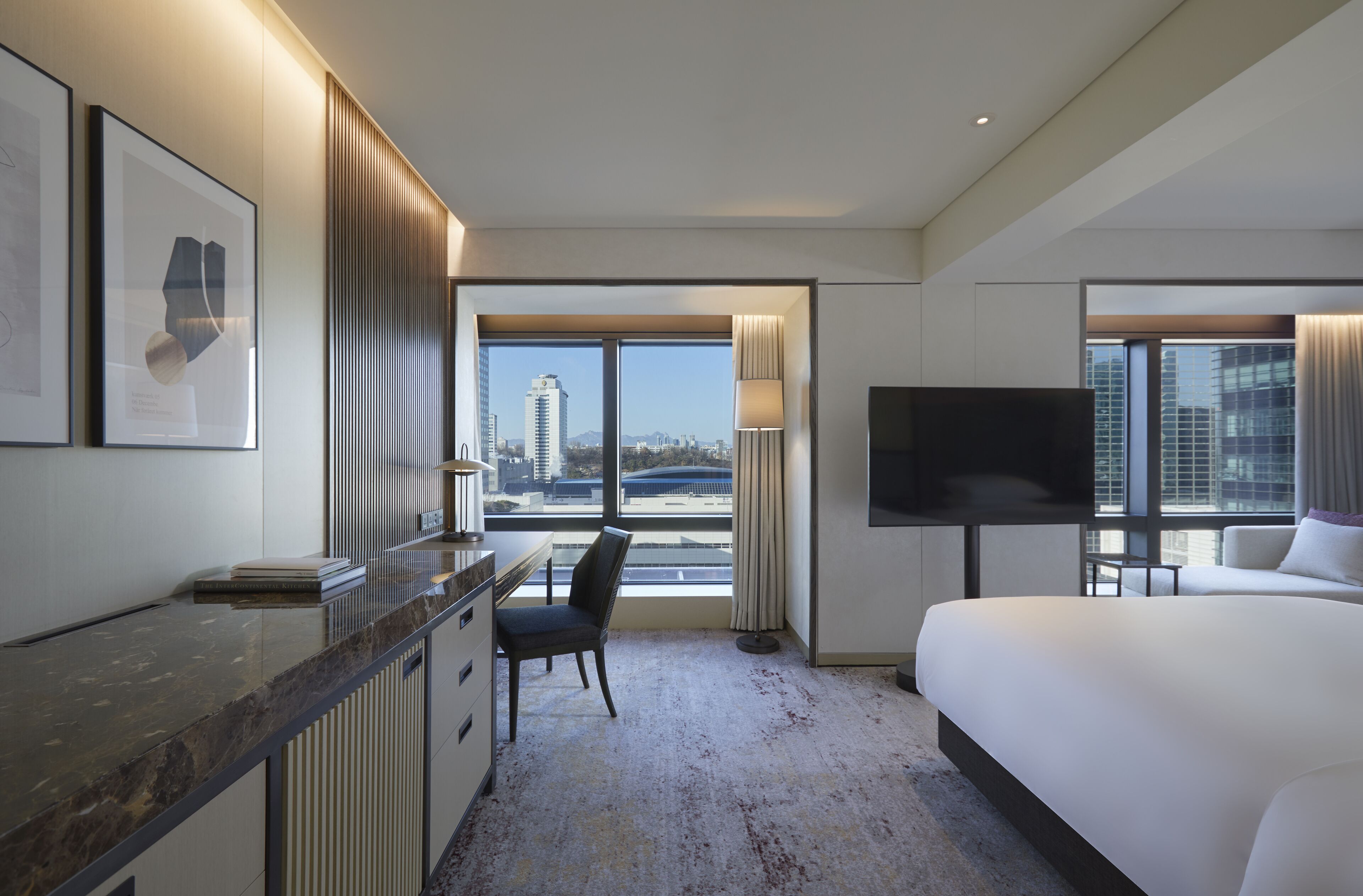 A hotel room with a bed, a desk, a chair, a TV, and large windows offering a city view.