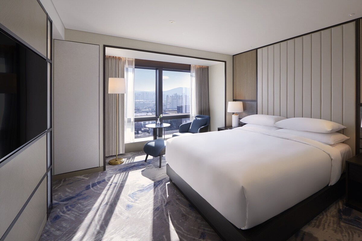 A modern hotel room with a large bed, two bedside tables, a TV, a window with curtains, and a city view.