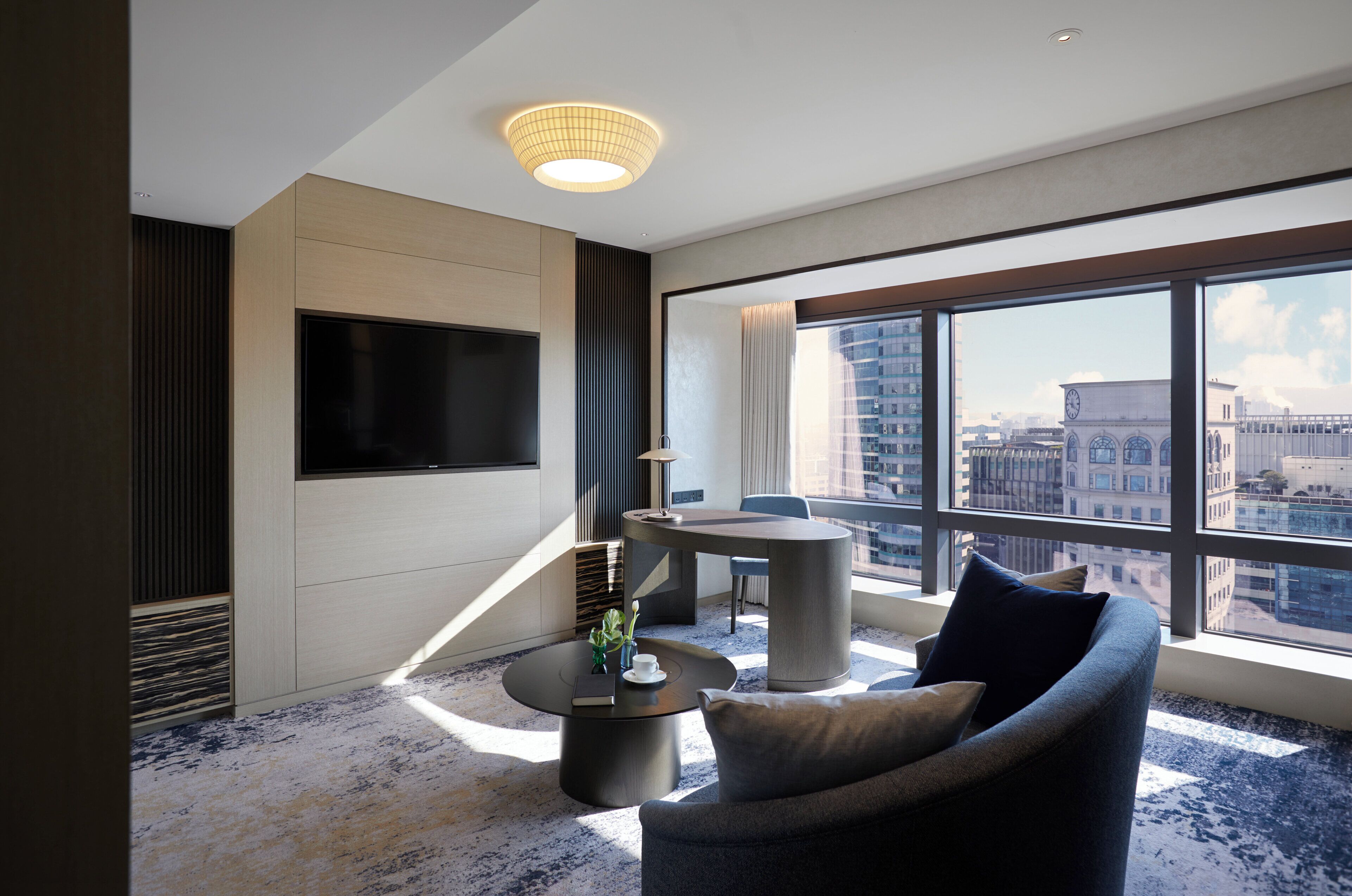 A modern hotel room with a large window, a flat-screen TV, a round coffee table, a sofa, and a small side table with a vase.